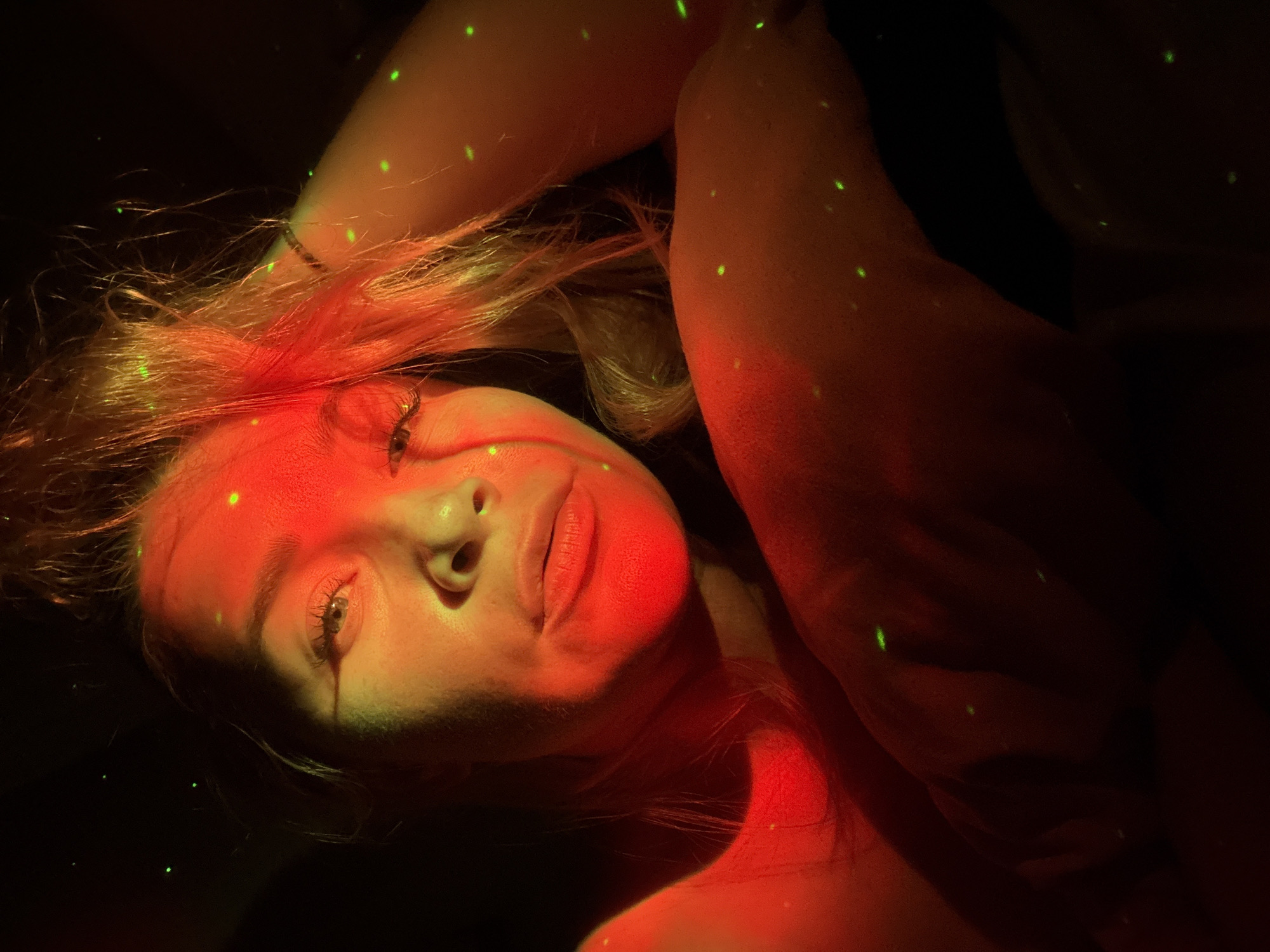 A close-up, chest-up portrait features a fair-skinned individual, identified as female, with light-colored hair, positioned in a dark, atmospheric indoor setting within the city of Veliky Novgorod, Russia. The subject's left eye is visible, appearing light, with long eyelashes. Her lips are full and slightly parted. She looks directly at the viewer. Her right arm is raised, with her hand near her head, subtly pushing back her hair. A thin, dark bracelet is visible on her right wrist. The scene is illuminated primarily by a strong red light source casting a prominent reddish hue on her face, neck, and upper body. Numerous small, bright green laser-like dots are scattered across her skin, hair, and the dark, indistinct background, indicating an artificial light projection. The environment is dark and lacks specific discernible features beyond the subject and the unique lighting effects.