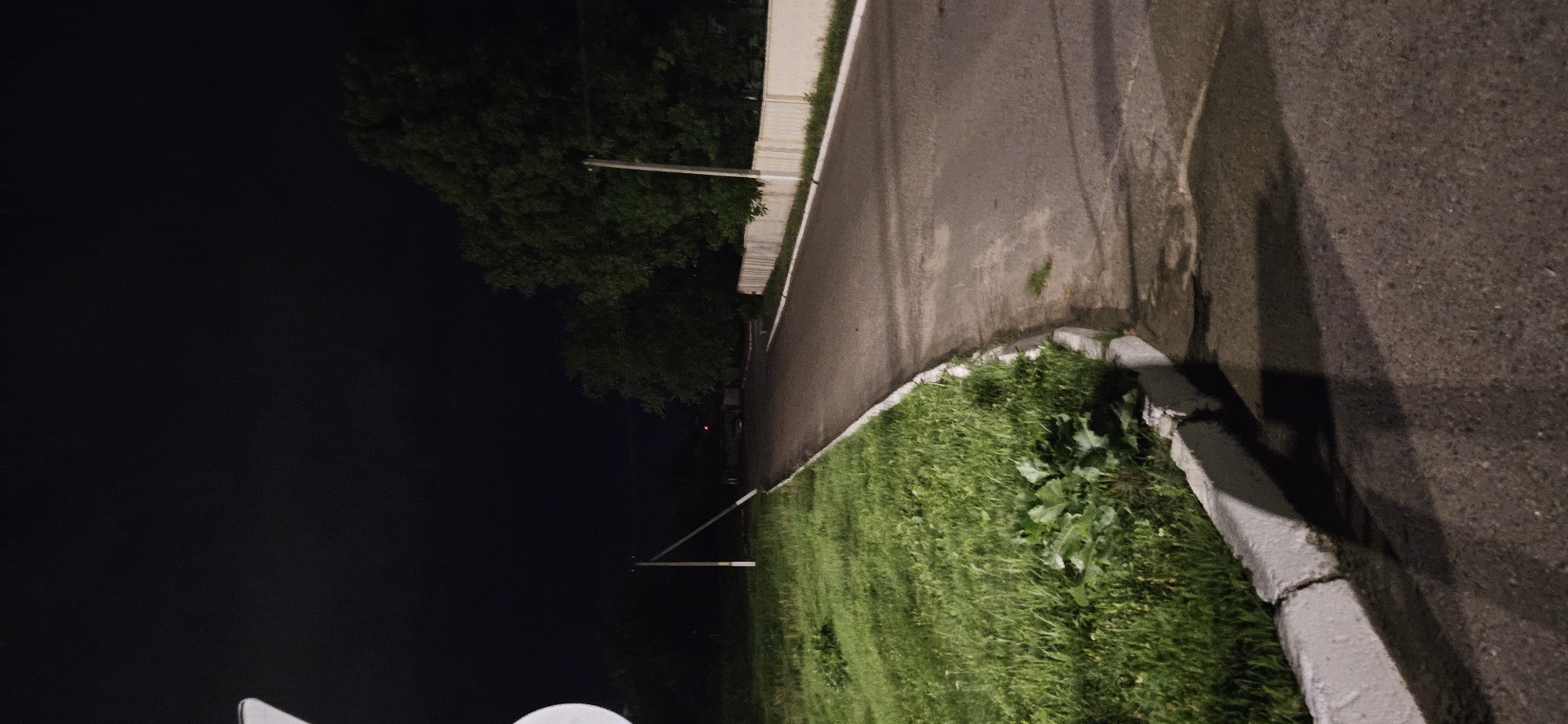 The image shows an asphalt road at night, bordered by a grassy area and a concrete curb on the left, and a white fence and trees on the right. The road curves away into the darkness. A utility pole is visible on the left side of the road. The sky is dark and overcast. It is unclear if this image is related to Vitsebsk, Belarus.
