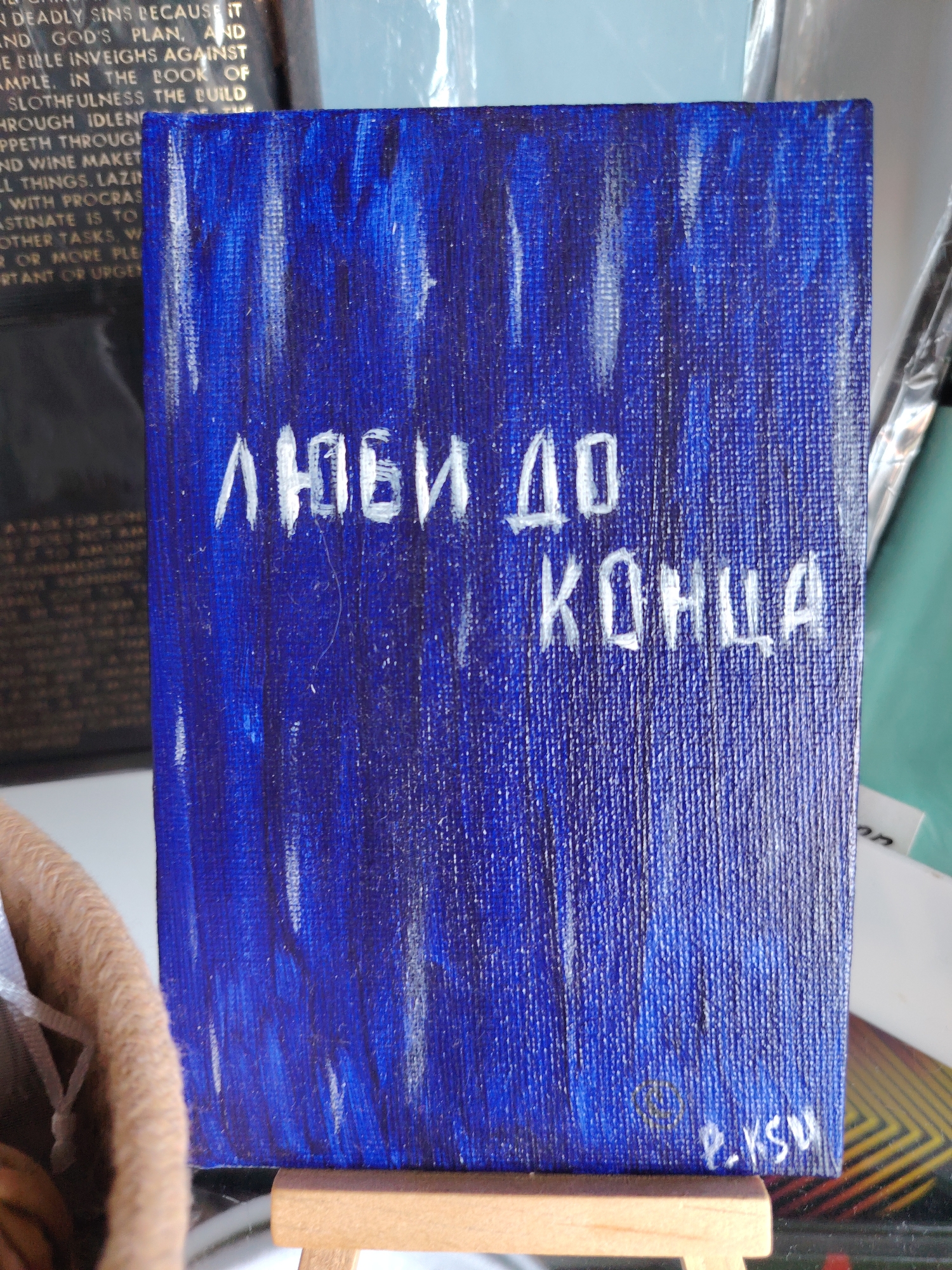 The image shows a small, rectangular painting with a dark blue background and white text. The text is written in Cyrillic and reads "ЛЮБИ ДО КОНЦА", which translates to "Love Until the End". The painting is displayed on a small wooden easel. To the left of the painting, a portion of a brown woven basket is visible, with a white ribbon tied around it. In the background, to the left of the painting, a blurred image of a book with gold lettering is partially visible. The overall scene appears to be an indoor setting, possibly a display or a craft fair, but the specific location in Konakovo, Russia, is not evident from the visual information provided.