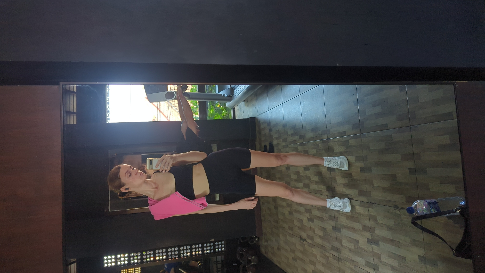 A slender, fair-skinned woman with auburn hair pulled back stands in what appears to be a gym, taking a mirror selfie. She is wearing a black sports bra, black bike shorts, and white athletic shoes. A pink towel is draped over her left shoulder. In her right hand, she holds a smartphone, also with a pink case.
The gym has a wooden tile floor and dark wood-paneled walls. To the left, gym equipment such as weight plates and kettlebells are visible. In the background, a treadmill is present, and through a window, greenery and a glimpse of buildings can be seen, suggesting an outdoor or semi-outdoor setting. A bottle of water and a dark purse are on the floor near the woman's feet. The image was captured in Mengwi, Indonesia.