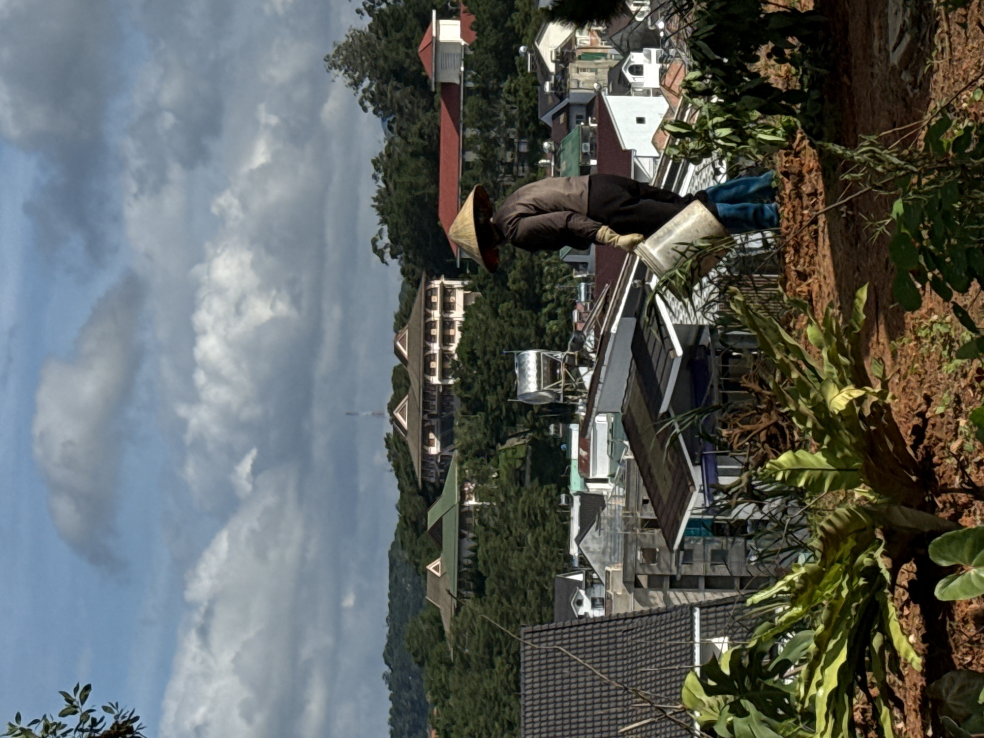 In the city of Lâm Viên - Đà Lạt, Vietnam, a person wearing a conical hat and work clothes stands on a hillside, holding a bucket and appearing to be watering plants. The person is dressed in a dark long-sleeved shirt, black pants, and blue rubber boots. They are also wearing gloves. The foreground features lush green foliage and exposed brown soil, indicating an outdoor garden or agricultural setting. In the background, a collection of buildings with various architectural styles, including houses and larger structures with red and white roofs, are nestled amongst dense green trees on rolling hills. The sky above is partly cloudy, with large white clouds scattered across a pale blue expanse. The overall scene suggests a rural or semi-rural area within the city, characterized by its natural landscape and residential development.
