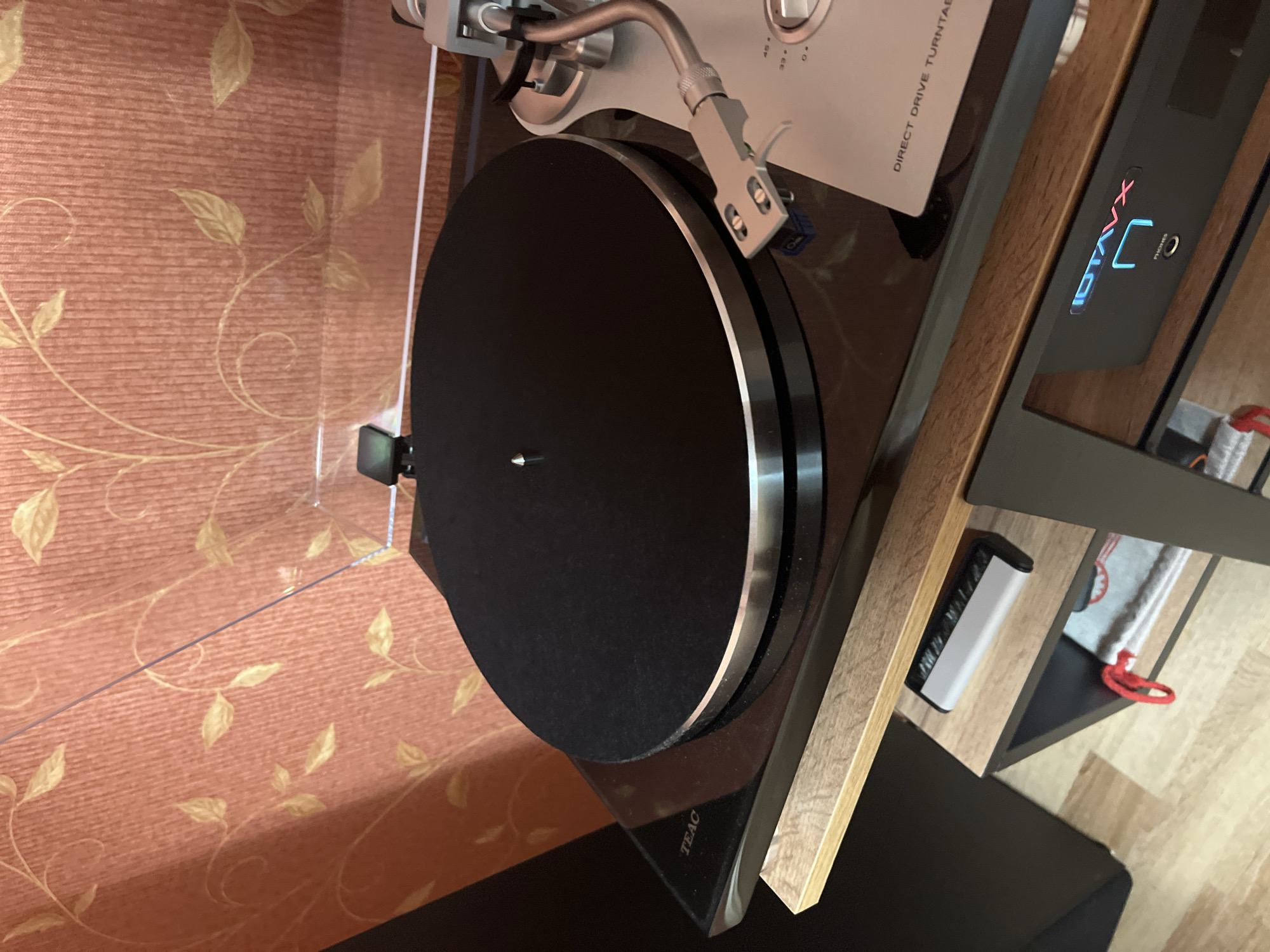 The image shows a close-up of a turntable with a black platter and silver accents. The turntable is placed on a glossy black surface, possibly the top of a component rack. To the left of the turntable, the "TEAC" logo is visible. A tonearm with a silver cartridge is positioned over the platter. The background features a textured wallpaper with a repeating pattern of light-colored leaves on a reddish-orange surface. Below the turntable, a wooden shelf is visible, on which a remote control and a piece of fabric with a red loop are placed. Further down, a black electronic device with illuminated "IDTAVX" and "PHONES" branding is visible. The flooring is a light-colored wood laminate. The scene appears to be an indoor setting, likely a living room or entertainment area. No people are present in the image. The location is identified as Ivanovo-Grushevsky, Russia.