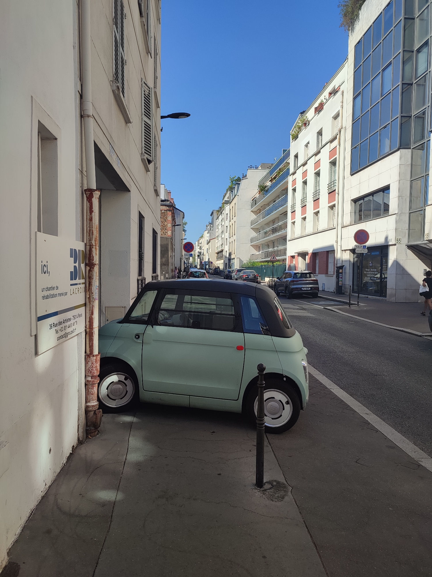 A light teal-colored, two-door electric car, identified as a Citroën Ami, is parked on the left side of a narrow, urban street. The car is positioned close to a white building, with its front wheels slightly turned towards the street. The building on the left has a white facade, a rusted downspout, and a sign that reads "ici, un chantier de réhabilitation mené par LACROIX".
The street is paved with asphalt and lined with parked cars and buildings on both sides. On the right side of the street, there are several multi-story buildings with balconies and windows. A blue sky is visible above.
Further down the street, traffic lights and road signs are present. A round red and white "No Parking" sign is visible on the right side of the street. Two pedestrians are walking away from the camera on the sidewalk on the right. The environment appears to be a typical European city street. The location is identified as Boulogne-Billancourt, France.