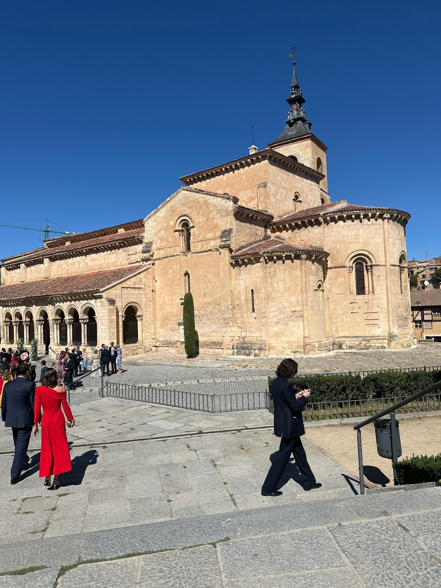 The image depicts the exterior of the Romanesque Cathedral of San Justo in Segovia, Spain. The sun is shining, casting strong shadows on the stone-paved ground and the light-colored stone facade of the church. The church features a prominent bell tower with a dark spire topped by a weather vane, and multiple arched windows and doorways. In the foreground, several people are walking. On the left, a couple in formal attire, a man in a dark suit and a woman in a vibrant red dress, are walking away from the camera, towards the church. Further back, a group of people, also dressed formally, are gathered near the entrance of the church. On the right side of the image, a person in a dark suit and striped pants is walking down a set of stone steps, looking at their phone. The environment includes a low metal fence and manicured green bushes.