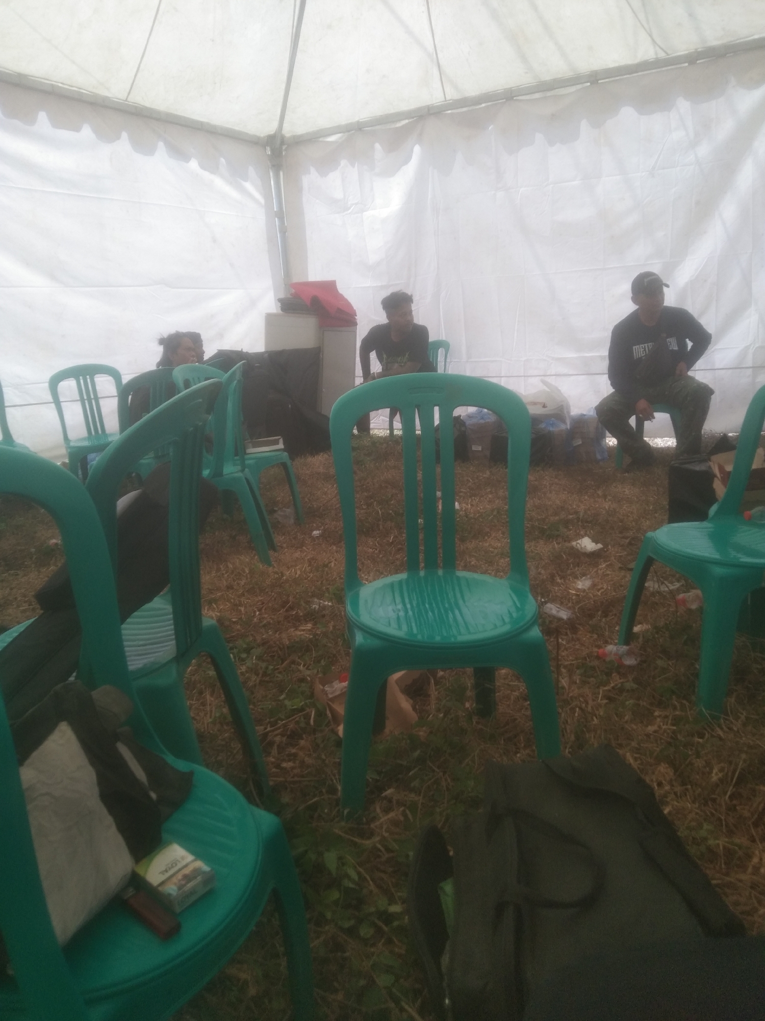 In Kecamatan Haurgeulis, Indonesia, a group of individuals are gathered inside a white tent. The ground is covered with dry grass and scattered debris. Several teal plastic chairs are arranged throughout the space.
In the background, three men are visible. One man, wearing a black long-sleeved shirt with writing and camouflage pants, is sitting on a chair, looking to his left. Another man, in a black t-shirt, is seated on a chair in the mid-ground, looking forward. To his left, a third man is partially visible, also seated.
In the foreground, several more teal plastic chairs are present. One chair has a pack of cigarettes and a lighter placed on its seat, along with a light-colored cloth and dark fabric. Another chair in the far foreground has a dark bag on it. The overall impression is a casual gathering or waiting area within a temporary structure.