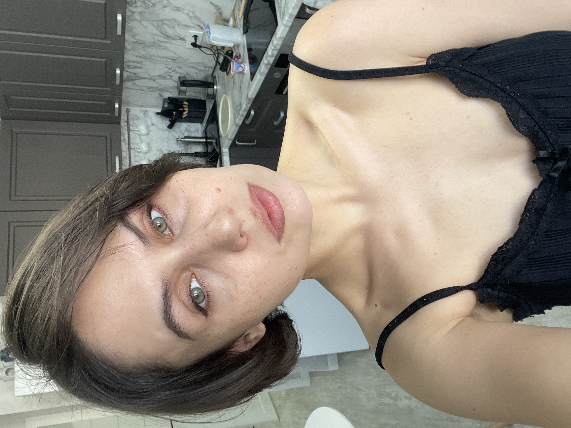 A woman with hazel eyes and freckles, wearing a black camisole, is photographed indoors. The background appears to be a kitchen, with dark grey cabinets, a marble countertop, and various kitchen appliances visible. The location in the city of Izhevsk, Russia is not discernible from the image.
