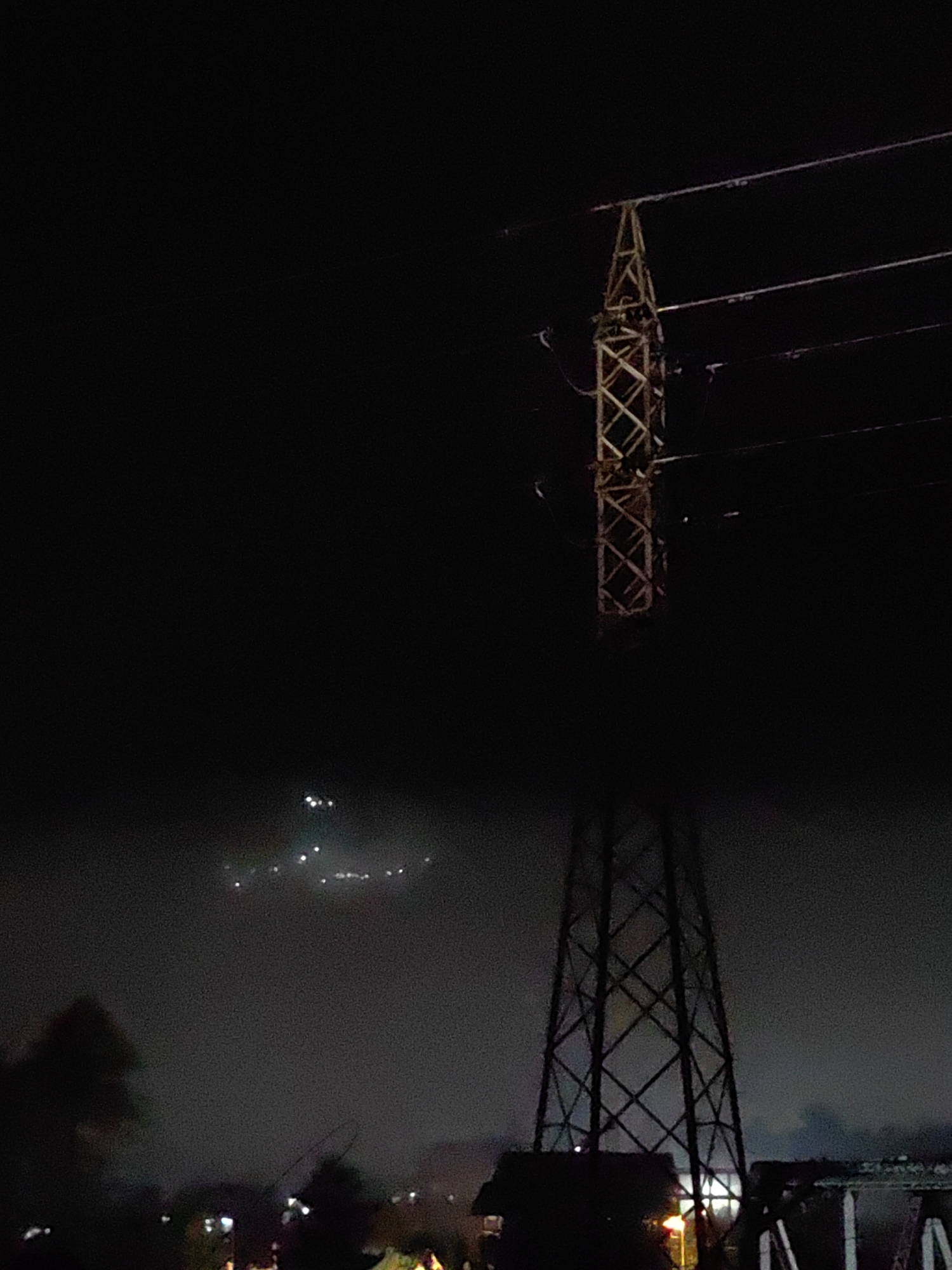 The image depicts a nighttime scene in Kulang, India, featuring two prominent electrical towers against a dark, hazy sky. The tower on the right is closer and more detailed, showing a latticework structure silhouetted against the dimly lit atmosphere. The tower on the left, further in the background, is partially obscured by mist or fog, with its upper portion visible against the sky.
Above the distant tower, a cluster of lights is visible, appearing as small, bright dots arranged in a scattered pattern. These lights suggest a settlement or activity occurring in the distance, further enhancing the sense of a rural or semi-urban setting at night. The overall mood is somewhat mysterious and atmospheric, amplified by the low visibility and the industrial presence of the electrical towers.
The foreground elements are largely obscured by darkness, with silhouettes of trees and possibly buildings visible at the bottom. The haze and darkness suggest a humid or foggy night, common in certain regions of India. The image captures a static moment, with no discernible activity or people. The time is clearly night due to the darkness and artificial lights. There is no visible text in the image.