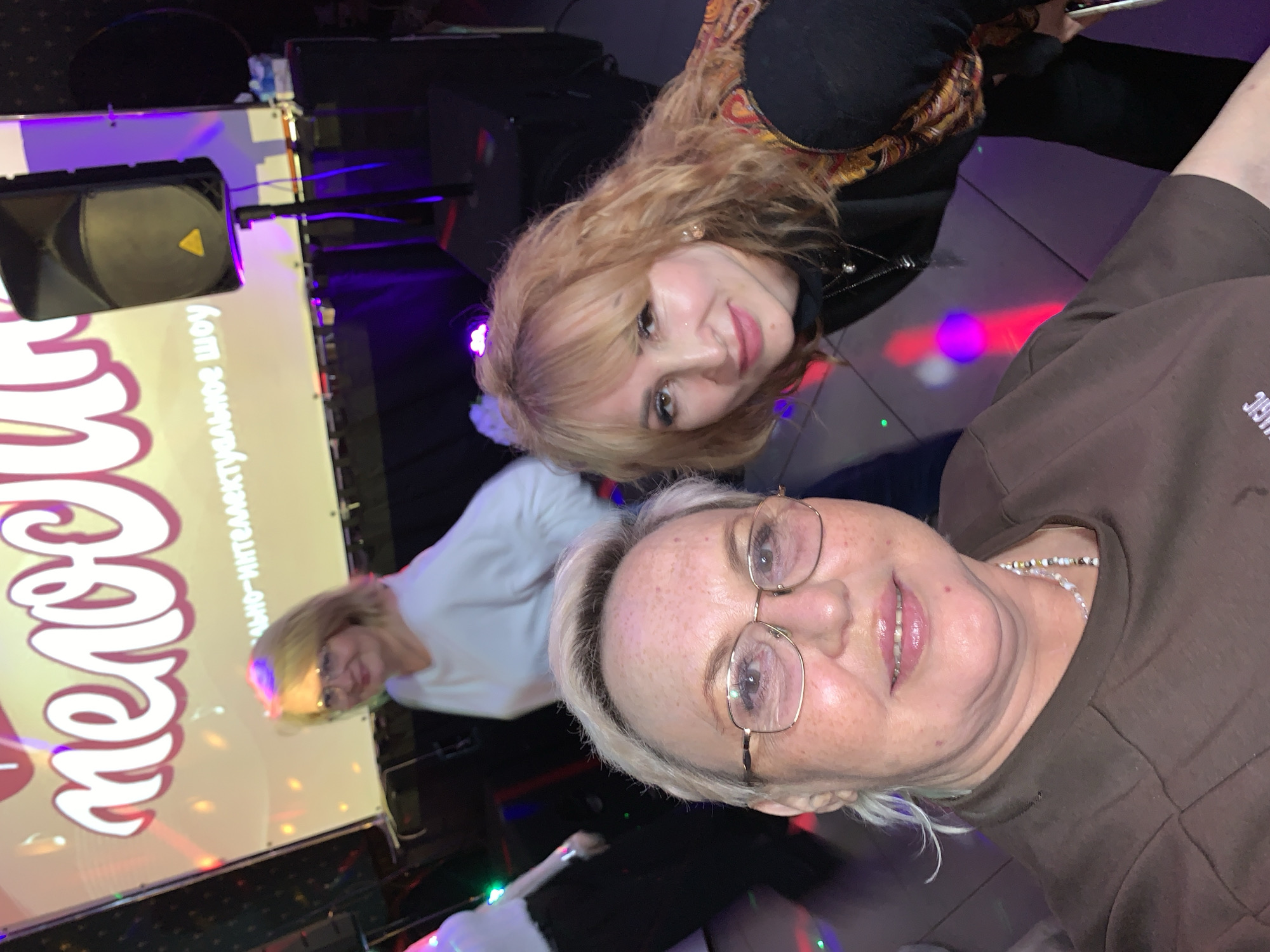 This image captures an indoor evening event featuring three women. In the foreground, an older woman with short, light-colored hair and glasses is shown from the chest up, smiling. She wears a brown long-sleeved top and a multi-colored beaded necklace. To her left and slightly behind, a younger woman with long, wavy blonde hair and a dark top with a patterned collar also smiles. Further back, a third woman with light hair and glasses, dressed in a white top, is visible, looking towards a large screen.
The setting is characterized by dim ambient light and vibrant, colorful spotlights casting red, purple, green, and blue reflections across the floor. In the background, a large screen or projection displays Cyrillic text. Prominently featured is the word "Телецкий" (Telecki) in a stylish white font with a pink outline. Below it, smaller white text reads "социально-интеллектуальное шоу" (social-intellectual show). A black speaker and a microphone stand are positioned near the screen, suggesting an entertainment or presentation setup. The expressions of the women convey a cheerful and engaged mood, indicative of a social gathering or performance in Chaikovsky, Russia.