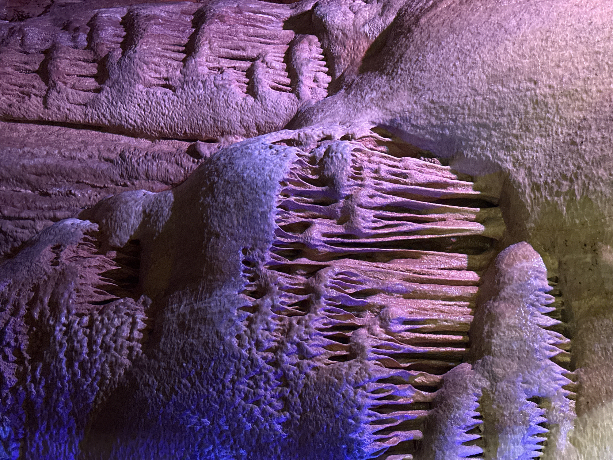 The image shows a close-up of cave formations in the Uhlovitsa cave, near Belo pole, Bulgaria. The formations include stalactites and flowstones, displaying textures of layered rock. Lighting highlights the structures with a combination of blue and pink hues.