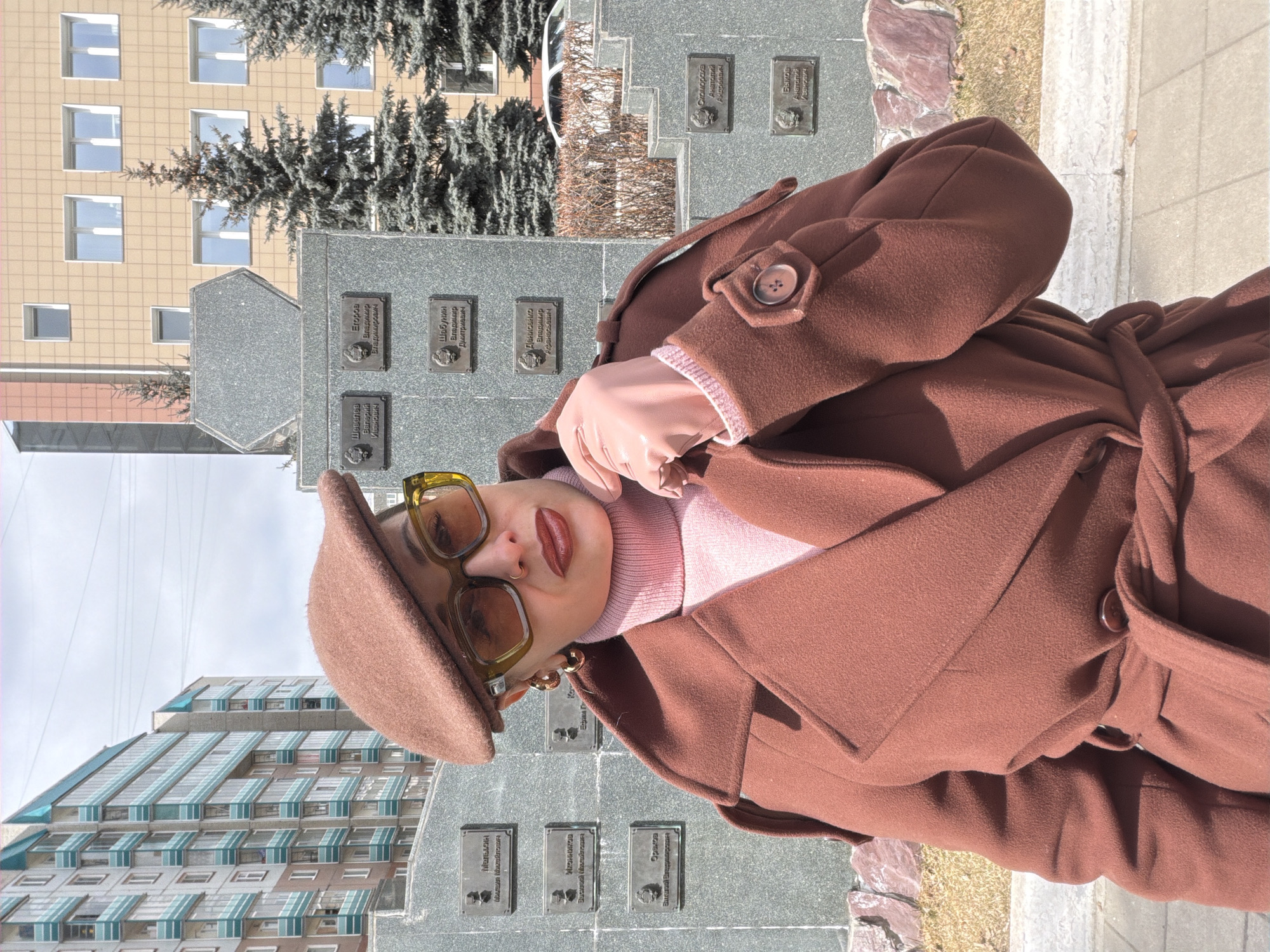 A stylish individual is posing for a portrait outdoors in what appears to be an urban setting, likely in Krasnoyarsk, Russia, given the contextual information and visible text. The person is wearing a fashionable brown wool coat, a pink turtleneck sweater, light pink gloves, a matching brown beret, and large, square-framed light brown sunglasses. They sport dark red lipstick and gold hoop earrings, conveying a composed and confident demeanor.
The background features a large, grey, granite-like wall or monument adorned with numerous bronze-colored plaques. These plaques bear etched Russian names, indicating they are likely memorial or commemorative markers. Visible names include "Шевелев Валерий Владимирович," "Егоров Владимир Иванович," "Шабунин Владимир Дмитриевич," "Денисенко Владимир Афанасьевич," "Скоморока Анатолий Андреевич," and "Быков Анатолий Иванович," among others.
Behind the monument, there are several tall coniferous trees and two distinct buildings. One building is a light beige or sand color with multiple rectangular windows. Another, partially visible, exhibits a unique architectural style with a sloped roof and many windows accented with light blue or green awnings/shades. The scene is illuminated by bright daylight, suggesting clear weather. The clothing worn by the person indicates a cool or mild climate.