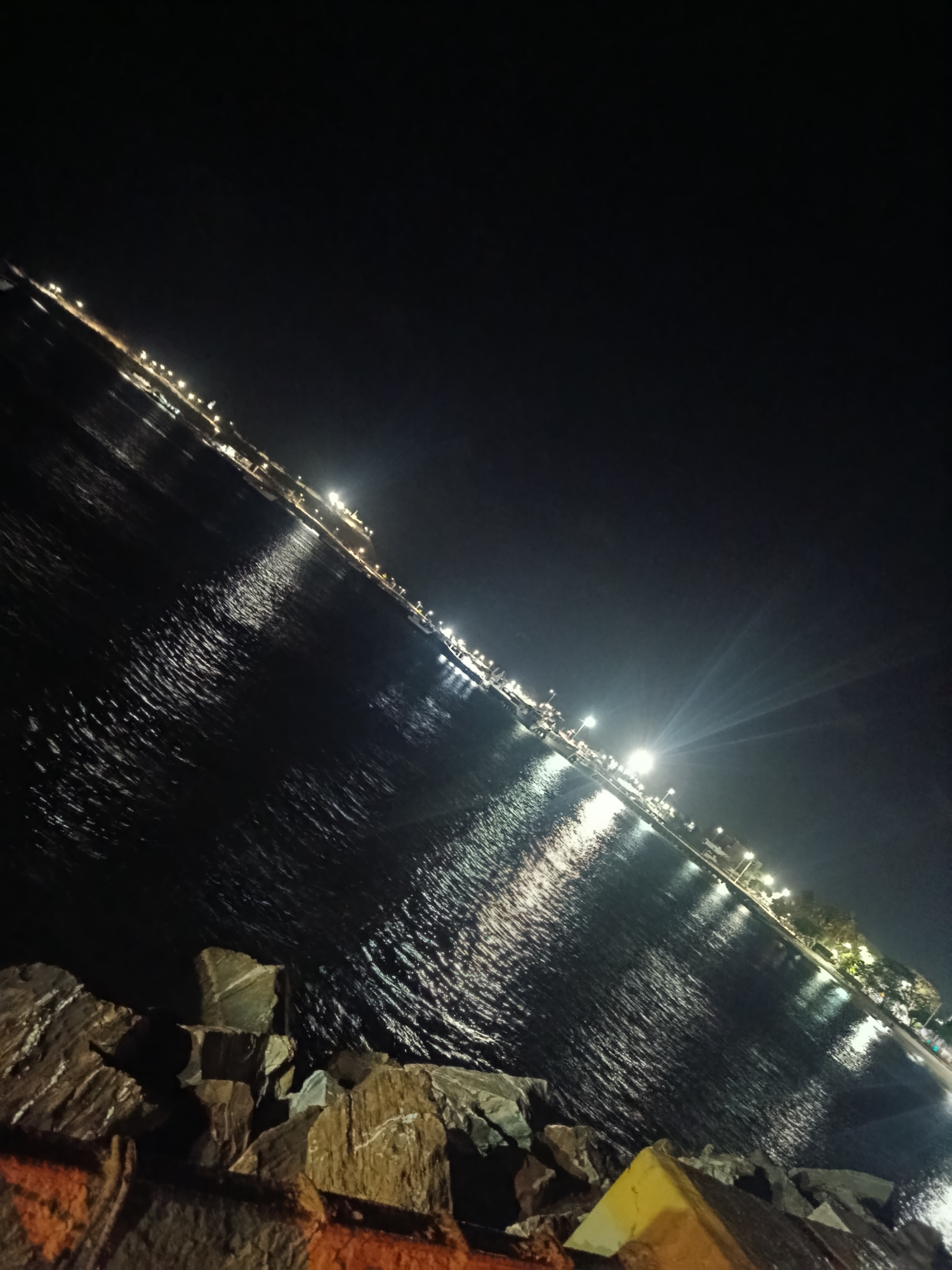 At night, the waterfront of Juan Griego, Venezuela is illuminated by streetlights that reflect off the dark water. In the foreground, a pile of rough, dark-colored rocks forms a barrier. The sky above is black, with no visible stars or moon. The angle of the photograph is canted slightly to the right, giving the scene a tilted perspective.