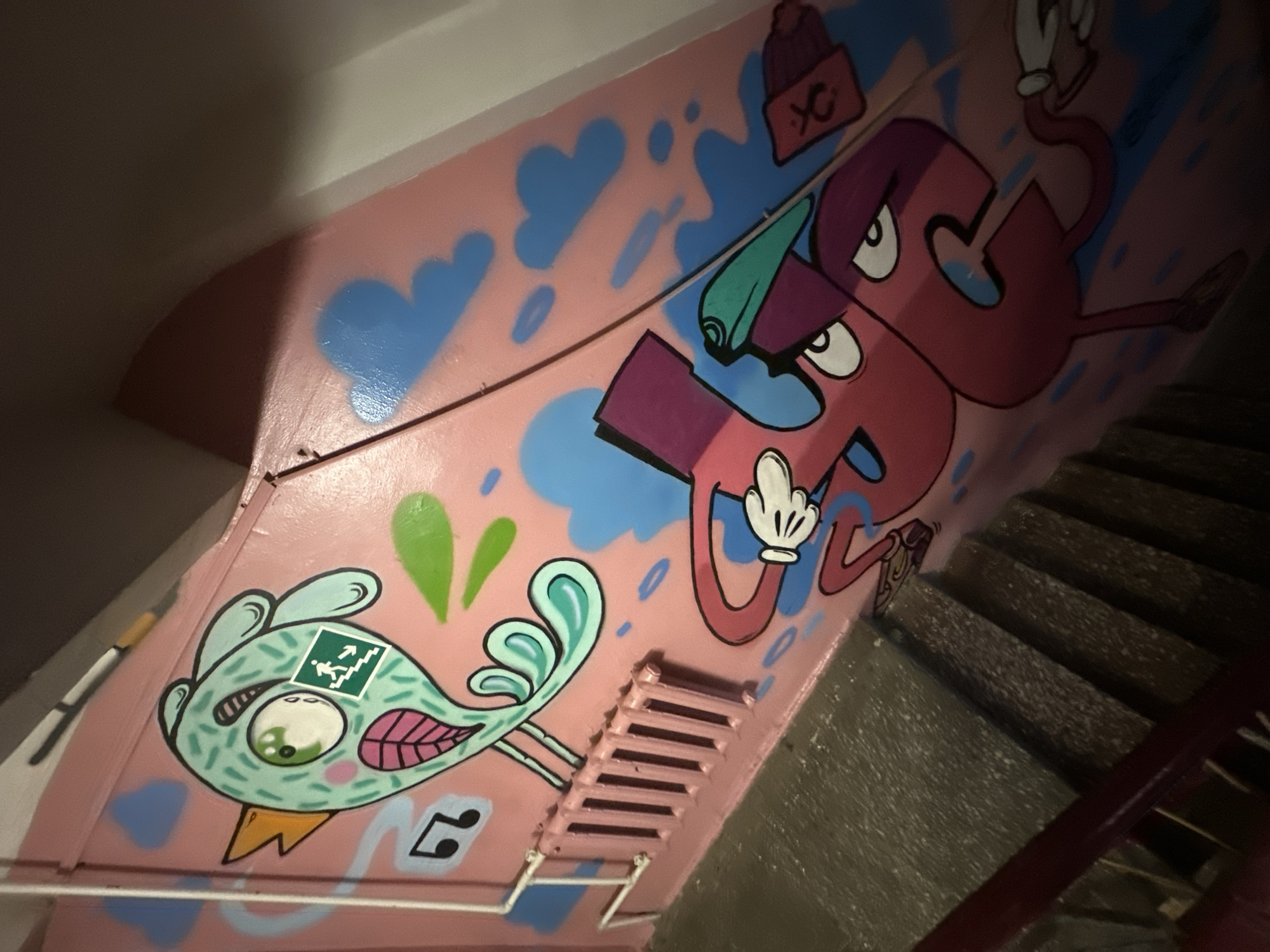 The image depicts a section of a stairwell decorated with graffiti. The walls are painted pink and adorned with cartoonish figures. On the left, a mint-green bird character with large eyes and a beak is depicted. A green square sign with a white silhouette of a person running down stairs is placed on the bird's cheek. To the right of the bird, a large, stylized pink character resembling a combination of numbers or letters, with arms, legs, and facial features, is drawn. This character wears a teal-colored beanie or hat. The background features blue cloud-like shapes and some musical notes near the bird. A pink radiator is visible on the wall below the bird. The stairs are made of speckled concrete, and a dark red handrail runs alongside them. The overall scene is likely a public or semi-public indoor space, possibly a residential building's stairwell, given the presence of the radiator and stairs. There is no specific visual information in the image that allows for identification of the location in Biysk, Russia.