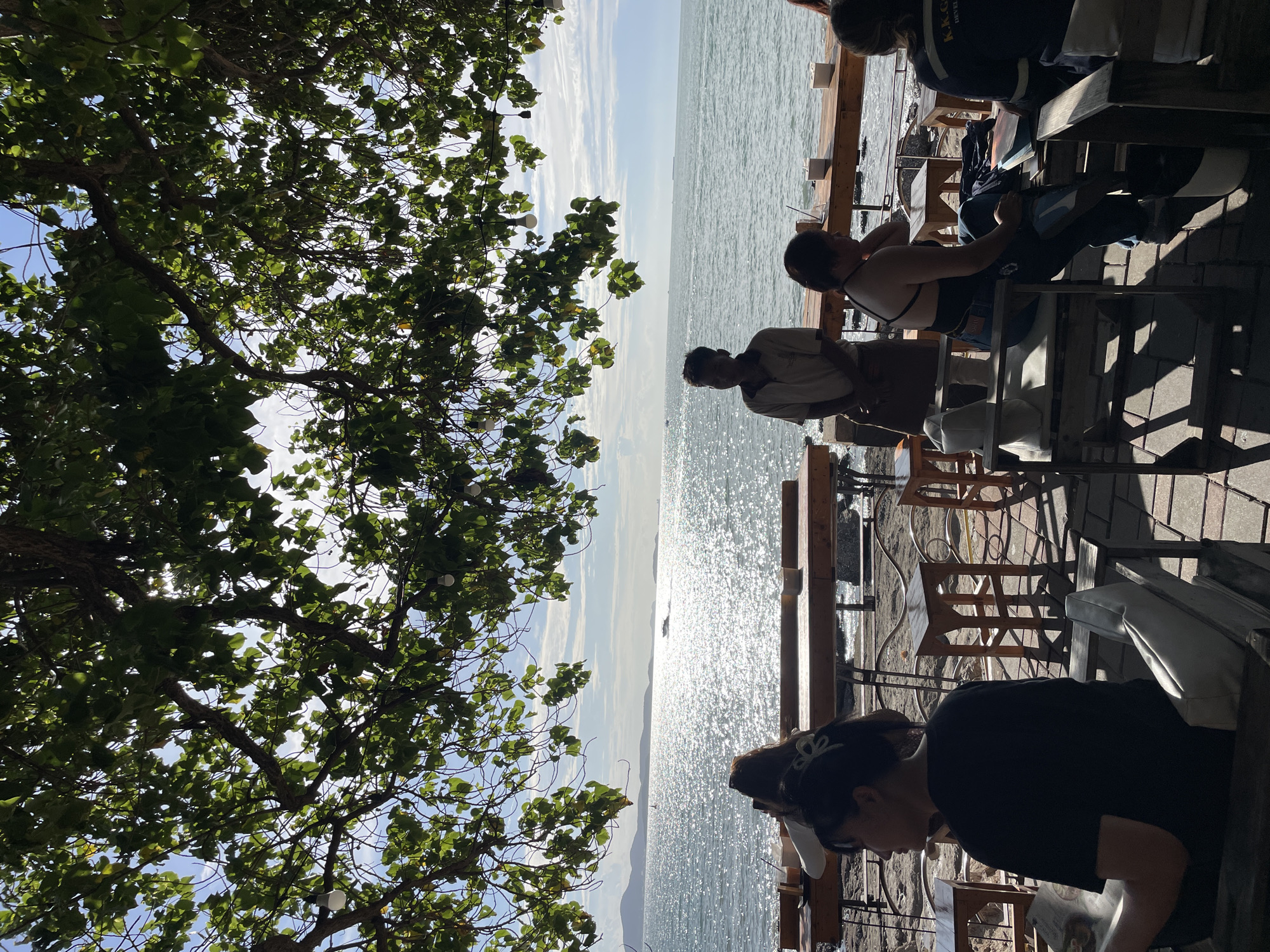 A photograph taken from beneath the dappled shade of a leafy tree canopy, looking out towards a sun-drenched ocean, likely in Pattaya City, Thailand. Several people are seated at outdoor tables, which are constructed from rough-hewn wood, positioned on a paved area. A man, possibly a server, stands near one of the tables, his attire suggesting a uniform. The ocean is a prominent feature, with sunlight glinting off its surface and a distant boat visible. The overall ambiance is that of a relaxed, seaside dining or lounging area.
