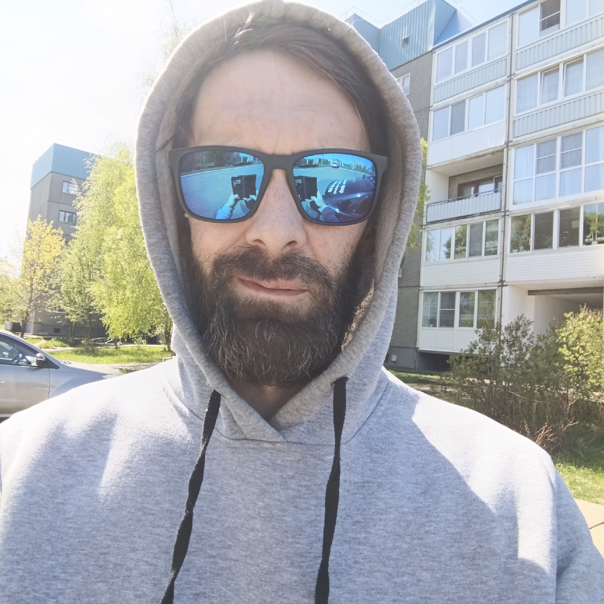 A man with a beard and sunglasses is wearing a grey hooded sweatshirt. The sunglasses reflect a street scene. The background shows residential buildings, trees, and a parked car. The location is Марковский, Russia.