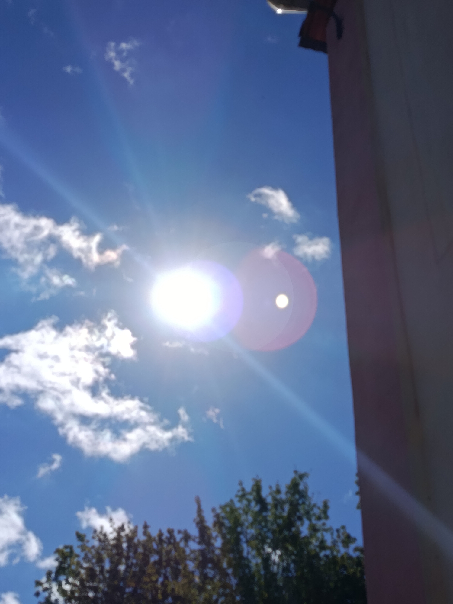 The image depicts a bright, sunny sky over Svietlahorsk, Belarus. The sun is intensely bright and appears as a large, white orb with a purple-blue halo, suggesting it is high in the sky. Sunbeams are visible radiating outwards from the sun, creating streaks of light across the blue expanse. A smaller, less intense circular lens flare is also visible to the right of the sun. Scattered white clouds, some fluffy and others more wispy, dot the sky.
To the right of the frame, a portion of a building is visible, its light-colored wall running vertically and obscuring part of the sky. At the very top right corner of the image, a dark, angled structure, possibly a roofline or overhang, is visible. In the bottom portion of the image, the tops of trees with green foliage are visible, indicating that the viewpoint is looking upwards from ground level or a slightly elevated position. The overall impression is of a clear, bright day.