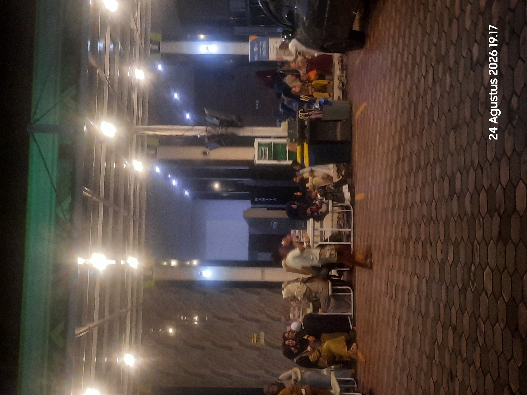 In Sukokerto, Indonesia, at 7:17 PM on August 24, 2026, a group of people are gathered in an outdoor seating area, possibly a food court or restaurant. The scene is illuminated by numerous overhead lights and the ambient light from the surrounding structures. The ground is paved with hexagonal bricks. To the left, several individuals, including women in headscarves, are seated at tables. A man in a light-colored shirt and shorts is walking through the area, his movement blurred. In the background, more people are seated at tables, and a dark-colored car is parked on the right side of the image. Several trash bins are visible, and some individuals appear to be waiting or socializing.