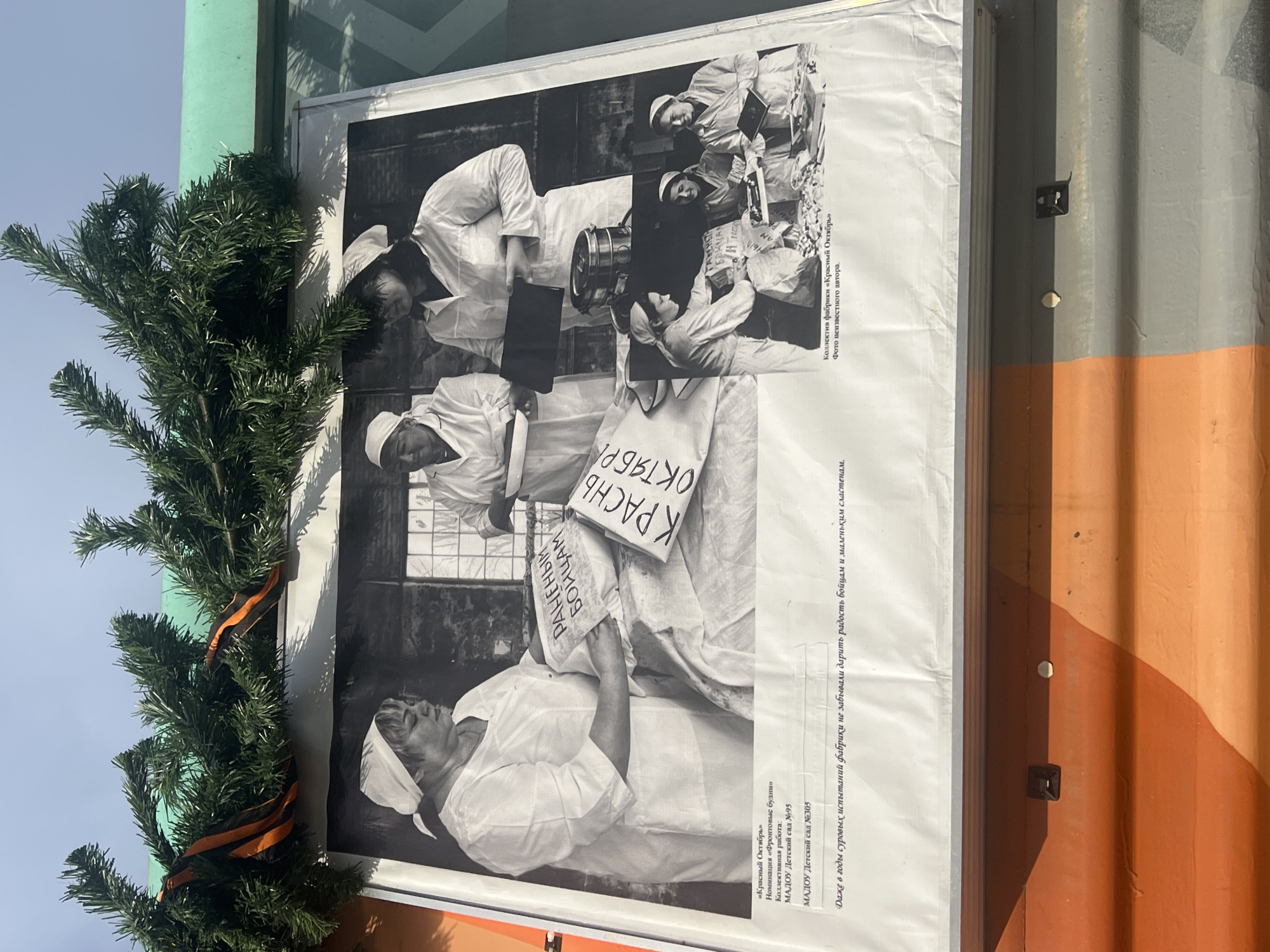 Here's a description of the image:
The image shows a poster displayed outdoors, possibly in Ufa, Russia, as indicated by the text on the poster. The poster features a black-and-white collage of photographs depicting scenes likely related to the World War II era, as implied by the text, which is written in Russian. The poster mentions "Krasny Oktyabr" (Red October), a nomination for "Front-line Days." The text also credits the "Front-line days" nomination to the collective work of MADOU kindergarten #95 and #305. The text on the poster reads "Krasny Oktyabr". The poster also mentions a photograph of the collective of the "Krasny Oktyabr" factory taken by an unknown author, stating that "Even in the hard years of trials, the factory did not forget to bring joy to the soldiers and little sweet lovers."
The photographs appear to depict women in white headscarves and uniforms, possibly factory workers or medical personnel. Some are shown holding signs with Russian text. The poster is attached to a building or container with orange and grey paint. To the left of the poster is an artificial green garland decorated with an orange and black ribbon. The photograph is taken in daylight.