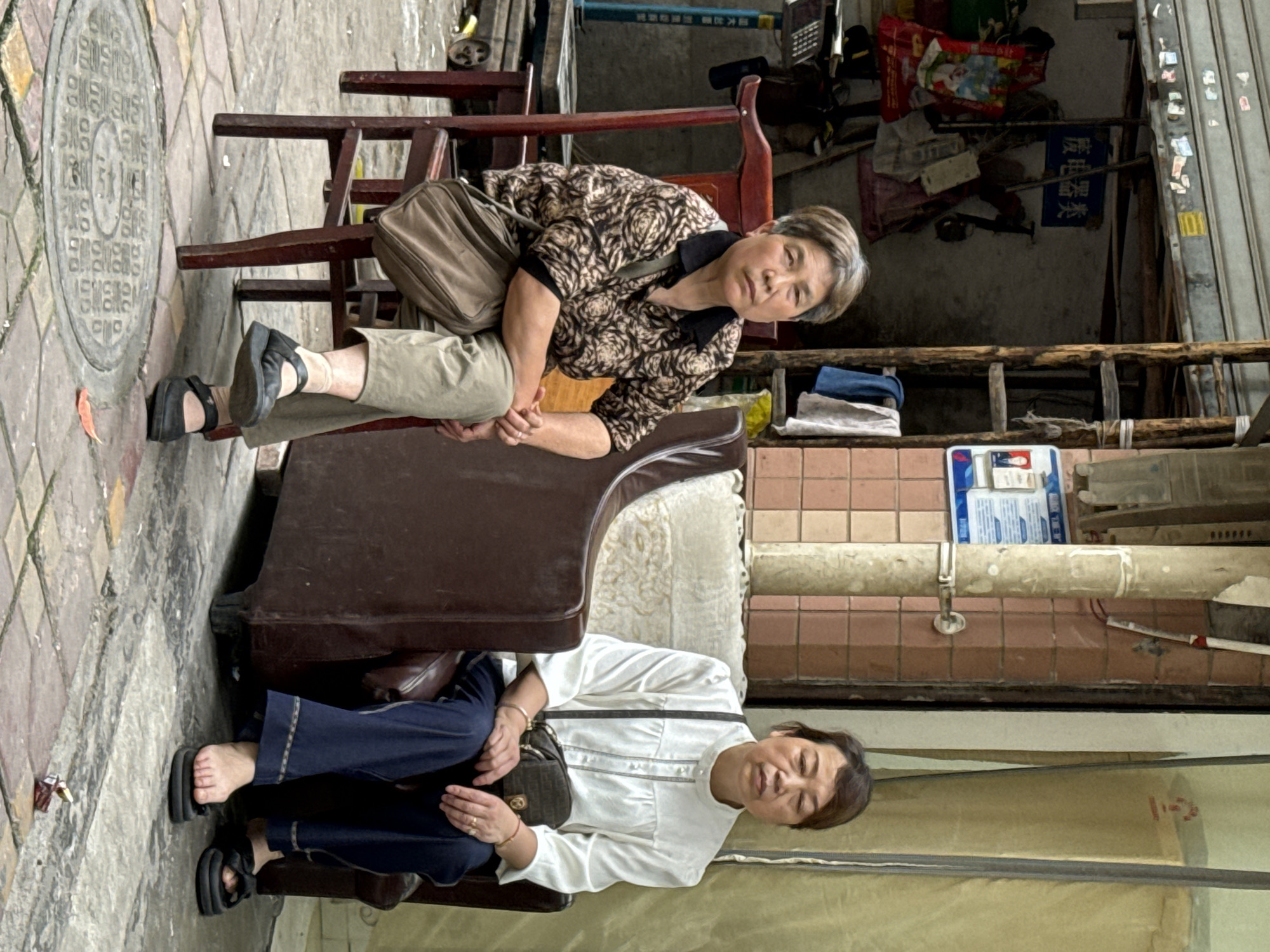 Two women are sitting in chairs in an alleyway. One woman is sitting in a red chair and is wearing a patterned shirt, tan pants, and black shoes. She has a brown bag on her shoulder. The other woman is sitting in a brown chair with a white cushion and is wearing a white shirt, blue pants, and black sandals. She also has a brown bag on her shoulder. The alleyway is narrow and cluttered. There are buildings on either side of the alleyway, and there is a sewer grate on the left side. The scene is in the city of Chongqing High-tech Zone, China.