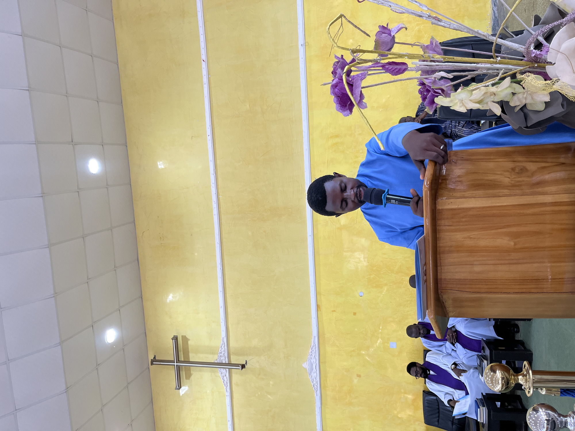 A man speaks at a wooden podium adorned with artificial flowers in a yellow-walled room with a cross displayed. Two men in white robes with purple stoles sit in the background. The location is possibly a church in Jos, Nigeria.