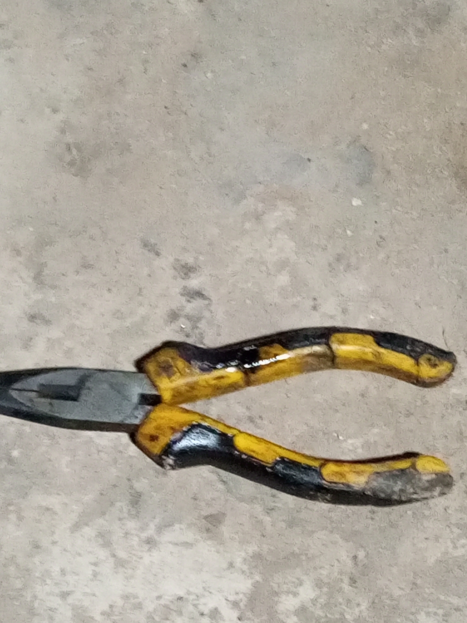 The image shows a pair of pliers lying on a textured, grey surface that appears to be concrete or asphalt. The pliers have yellow and black insulated handles. The metal jaws of the pliers are visible and are open. The background is out of focus and appears to be a continuation of the grey surface. There are no people or other objects clearly visible in the image. The lighting is somewhat flat, and the image quality is not very sharp, suggesting it may have been taken with a low-resolution camera or under poor lighting conditions. There is no information in the image that would indicate its specific location within Keling Panggau, Indonesia.
