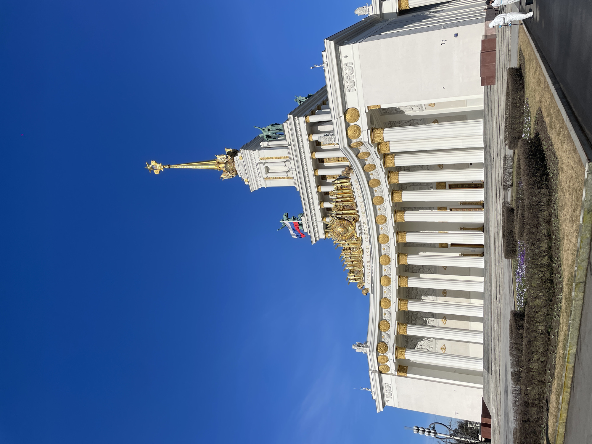 The image shows the facade of the Central Pavilion at the Exhibition of Achievements of National Economy (VDNKh) in Moscow, Russia. The architecture is characterized by white columns, gold detailing, statues, and a spire topped with a star. The Russian flag is visible atop the building. The sky is a clear blue. A groomed garden is visible to the right. Two people wearing coveralls are visible near the top edge of the image.