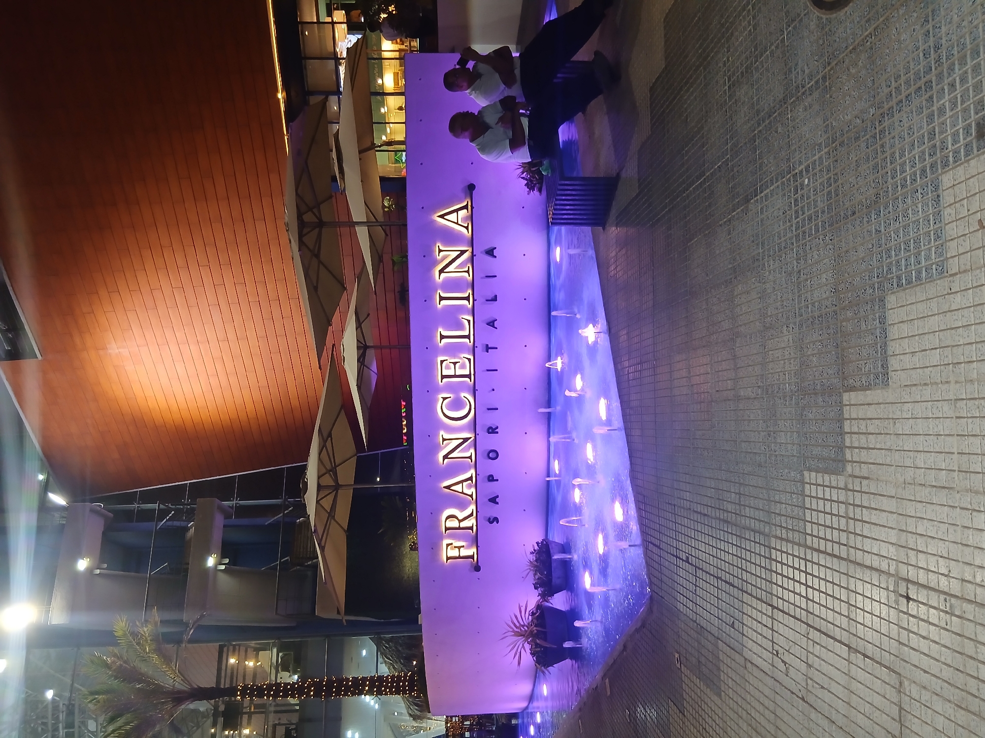 The image shows the "FRANCELINA Sapori Italia" restaurant in Porlamar, Venezuela. The restaurant's name is vertically displayed on a purple wall with a water feature below. Two people are sitting on a bench near the sign. The scene appears to be at night. The surrounding area includes tiled pavement, umbrellas, a palm tree, and other building structures.