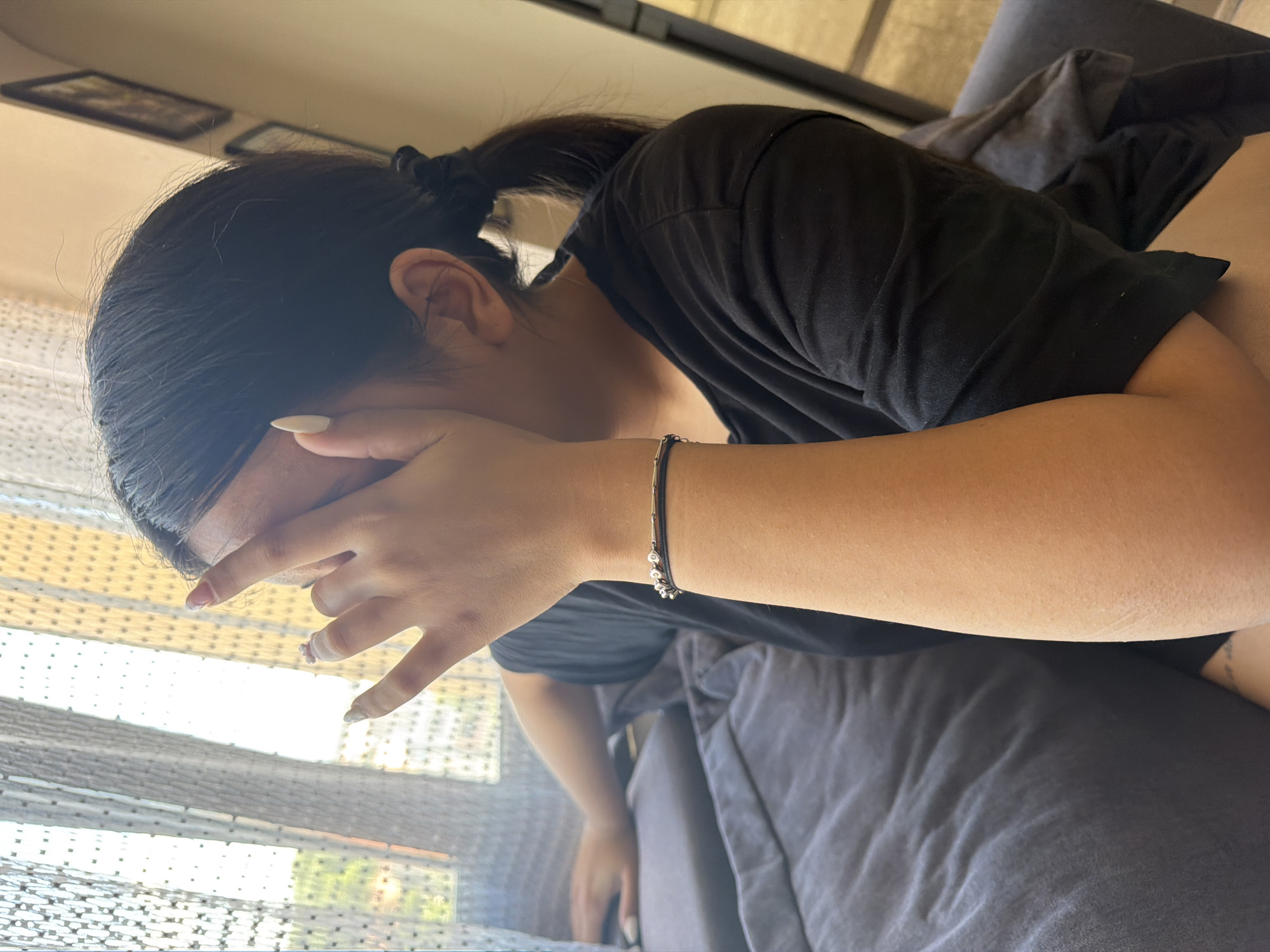 The image captures a close-up of a young woman's face and arm as she sits on a sofa. She is wearing a black t-shirt and has her hand covering her face, obscuring her eyes. A black bracelet with silver accents adorns her wrist. The background reveals a sheer curtain, through which sunlight filters, and a framed picture on the wall. A portion of a dark-colored sofa is visible in the foreground. The scene appears to be indoors, possibly a living room. The image does not provide information about the city of Shumen, Bulgaria, or any specific location within it.