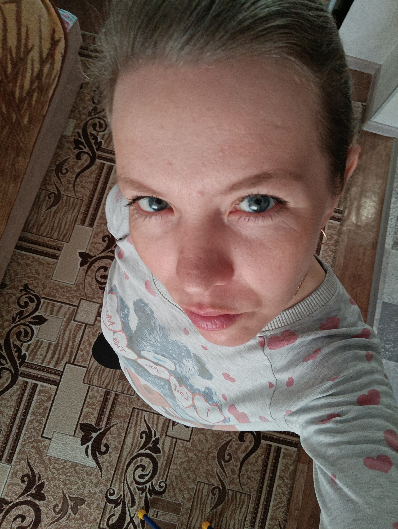 A high-angle selfie of a fair-skinned woman with blue eyes. Her hair is pulled back. She is wearing a gray long-sleeved shirt with pink hearts. The background shows a patterned floor. A section of a beige couch is partially visible in the upper left. The location is Chaikovsky, Russia.