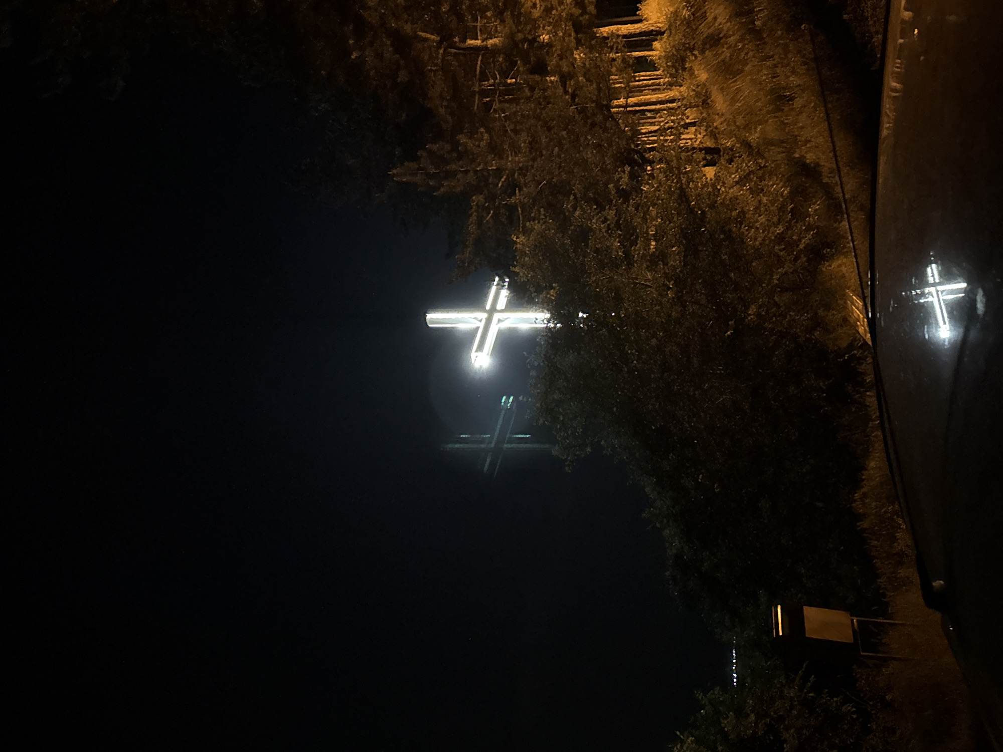 The image shows a nighttime scene in Blagoevgrad, Bulgaria. In the distance, two illuminated crosses are visible against a dark sky. The surrounding environment includes trees and vegetation on either side of the frame. A dark object, possibly a vehicle part, is present on the right side of the image, reflecting the light from the crosses. A sign is also visible near the bottom-right corner.