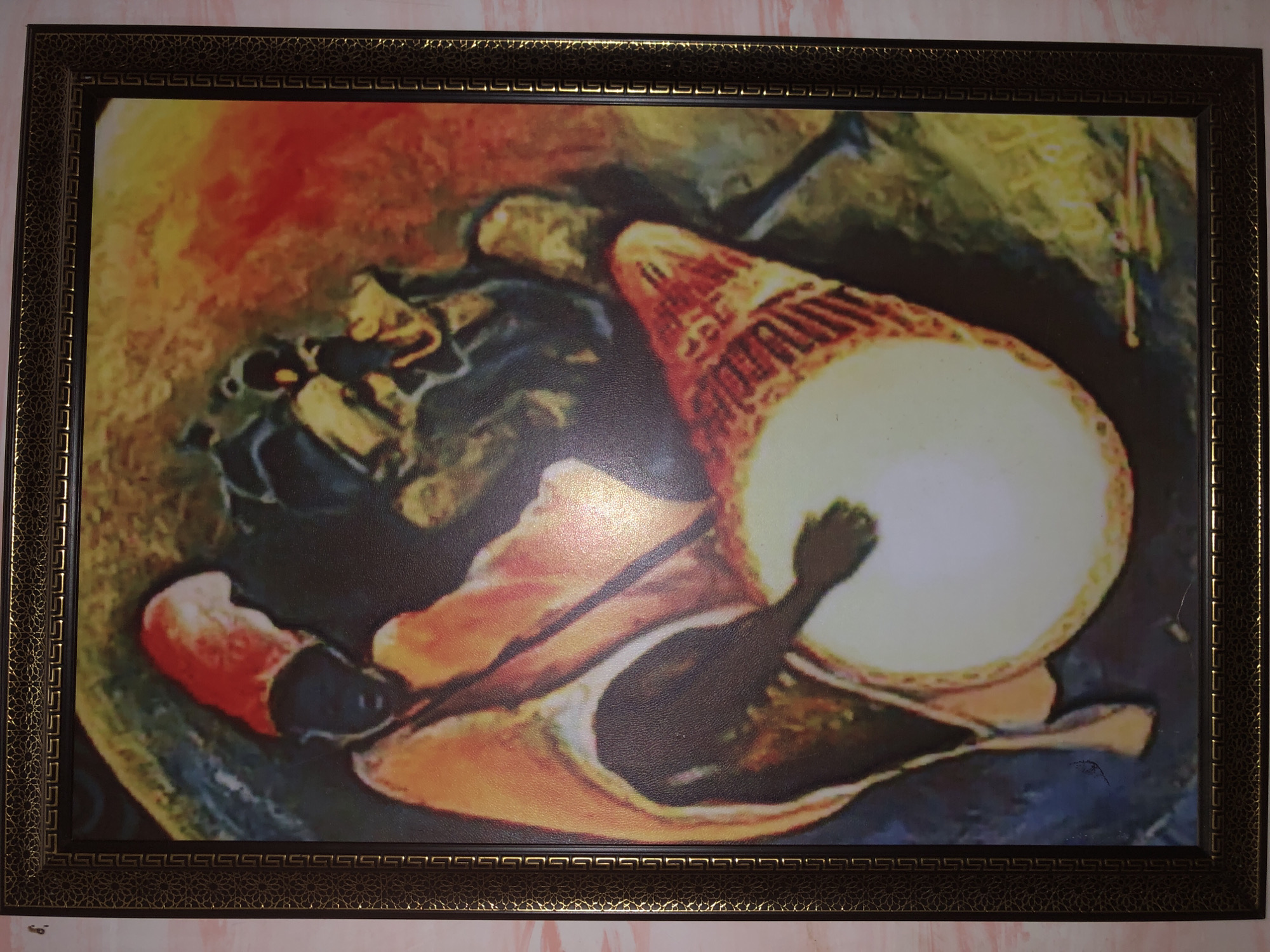 The image shows a framed artwork hanging on a wall. The artwork depicts a person playing a drum. The person wears a red hat and is rendered in dark hues. The drum is large and predominantly white with orange and red accents. The background features a blend of red, yellow, and dark colors. The frame is dark with intricate geometric patterns. The location is Unknown, Unknown.