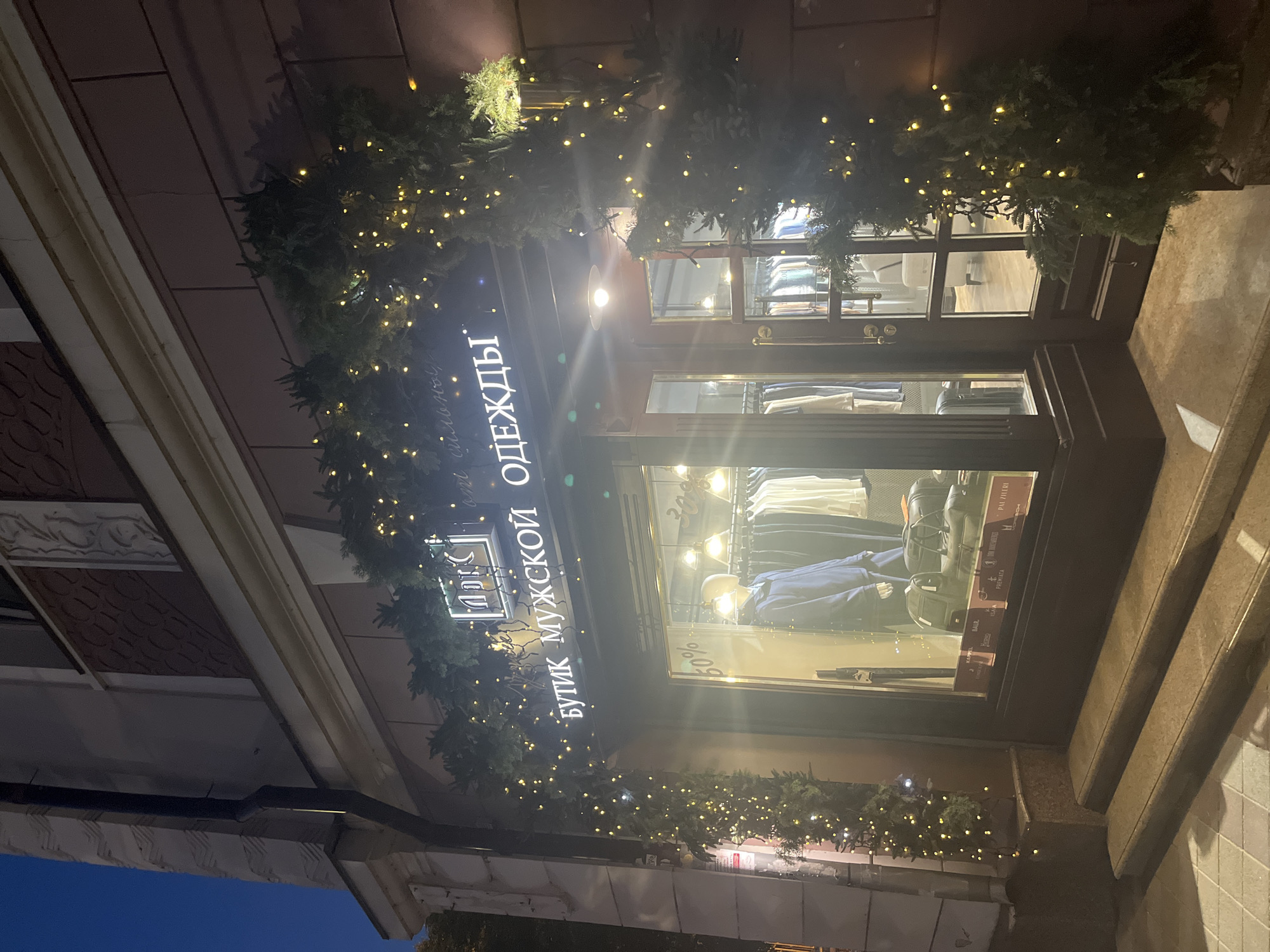 The image shows the facade of a store at night. The store is decorated with evergreen branches and twinkling lights, suggesting a holiday or festive season. The sign above the entrance reads "БУТИК МУЖСКОЙ ОДЕЖДЫ" which translates to "Men's Clothing Boutique." In the window display, mannequins are dressed in suits and there are shelves with folded clothing. Some text on the window indicates sales, with "30%" and "50%" visible. The overall atmosphere appears to be upscale and inviting, with the warm lights creating a cozy ambiance against the dark sky. The building appears to be made of brick and stone, with a decorative cornice. The presence of steps leading up to the entrance suggests a ground-level establishment. The location is identified as Krasnodar, Russia.