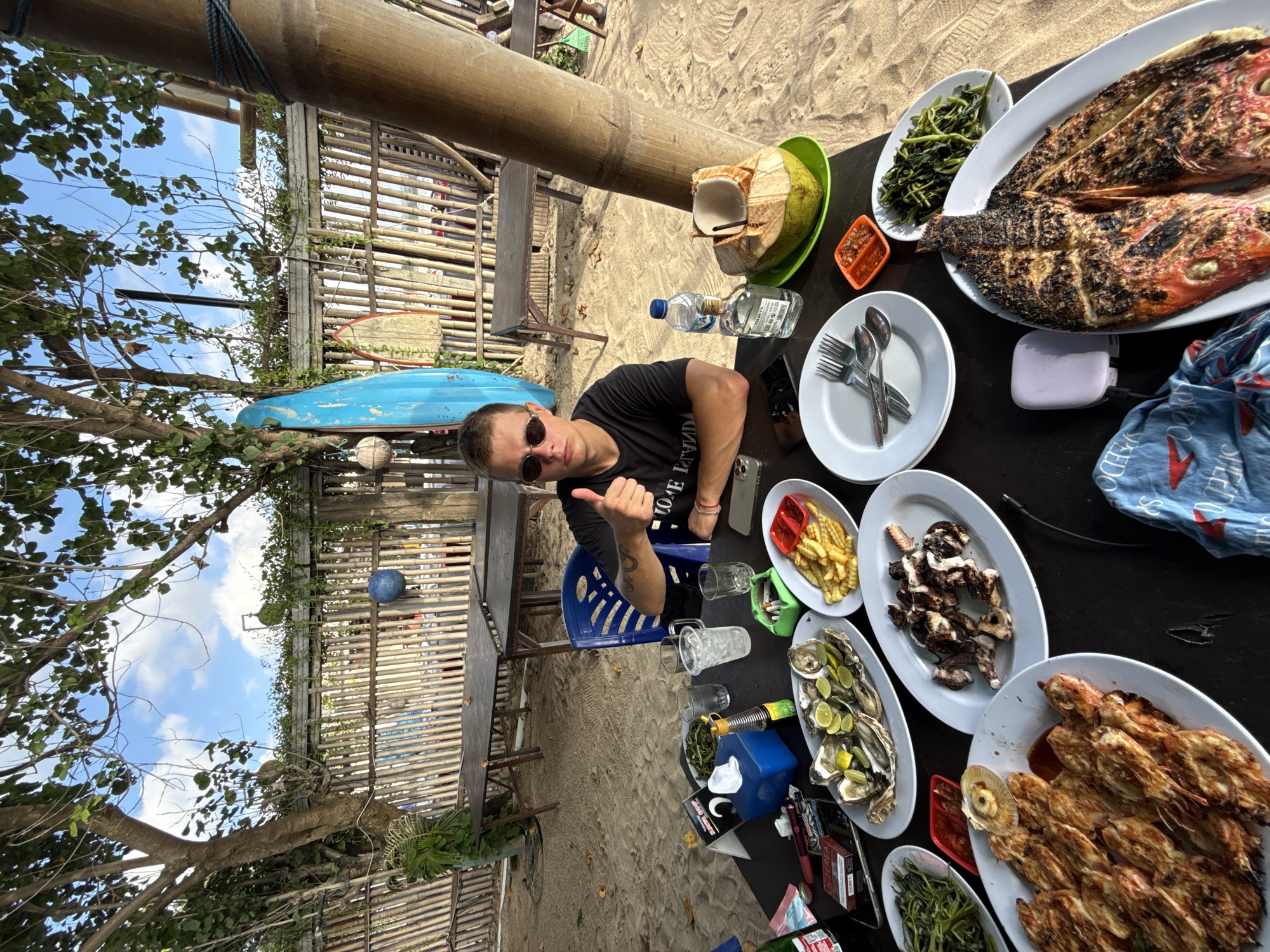 The image captures a dining scene in Kuta, Indonesia. A man wearing sunglasses and a black t-shirt gives a thumbs-up gesture, sitting at a black table laden with food on a beach restaurant. The table includes plates of grilled fish, grilled shrimp, oysters with lime wedges, grilled octopus, french fries, and greens. A coconut drink, bottled water, cutlery, and personal items like phones and a power bank are also present. A blue plastic chair is visible, and in the background, there is a bamboo structure, beach sand, and the sea. A tree branch stretches across the left side of the frame, partially obscuring the sky.