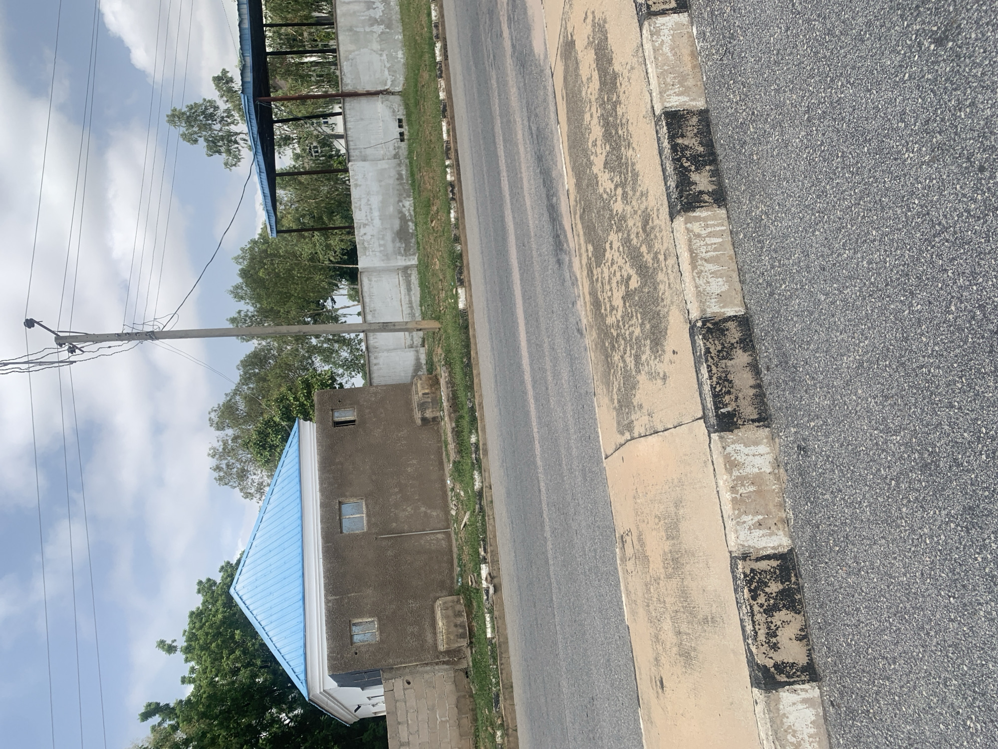 The image depicts a residential area on the outskirts of Jalingo, Nigeria, captured on a partly cloudy day. A street with a concrete curb and asphalt surface occupies the foreground. Beyond the street, a light brown, stucco-sided building with a bright blue, metal-sheeted gabled roof is visible. The building has three small, rectangular windows. To the left of this building, a section of a concrete block wall is present. Further back, a light grey, stucco-finished boundary wall runs along the property line. Behind this wall, a covered structure with a blue, metal roof supported by dark poles is situated. Lush green trees and vegetation surround the buildings. A concrete utility pole with electrical wires stretches across the top portion of the image. No people are visible in the scene. The overall impression is one of a developing suburban environment.