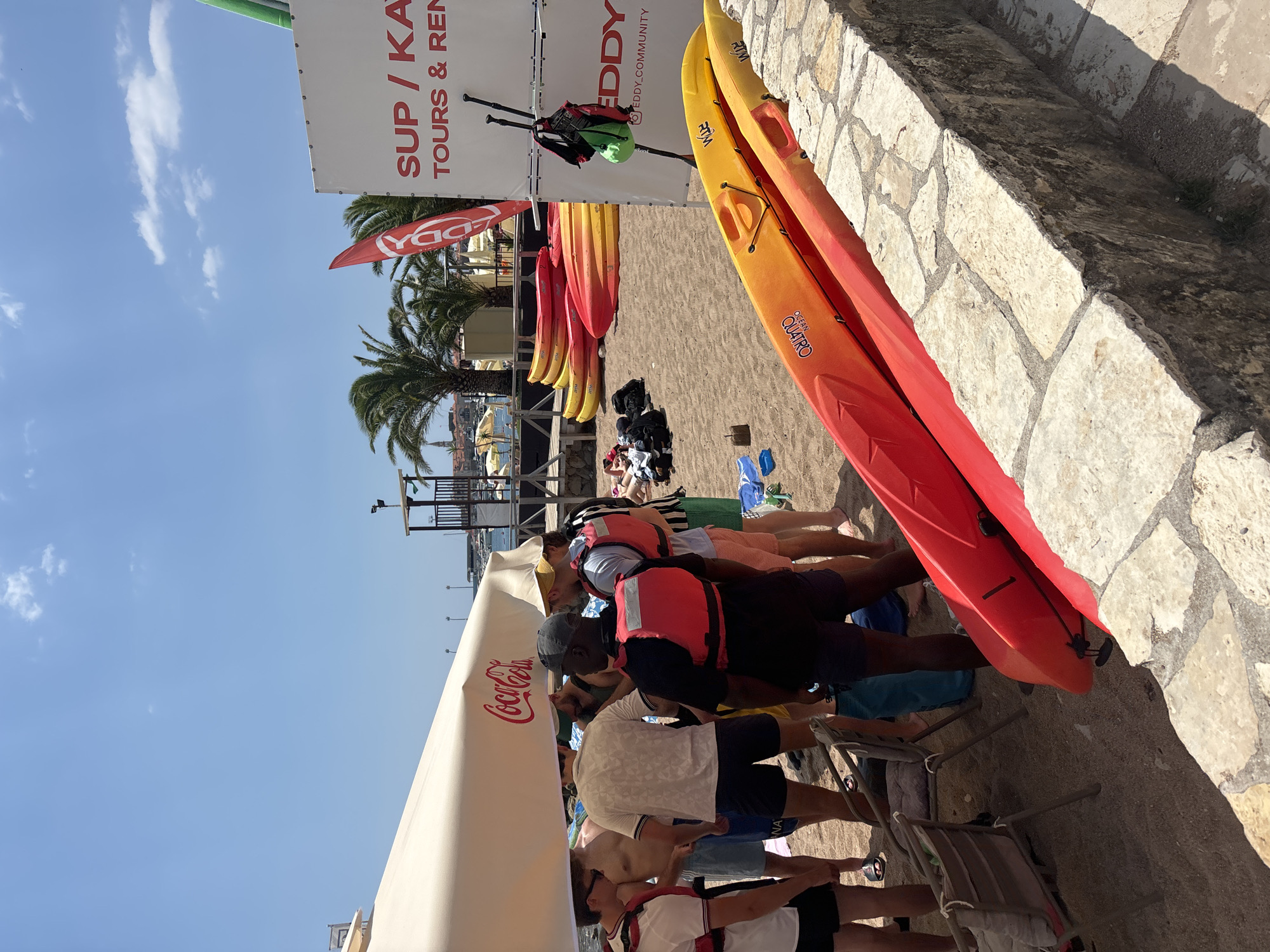 A group of young men stand clustered under a large beige umbrella on a sandy beach in Budva, Montenegro, with some wearing life vests, indicating a planned water activity. In the background, palm trees line the coast, and buildings with red roofs suggest a Mediterranean setting. Several kayaks, some red and orange, are stacked on the sand and near a stone wall, hinting at a kayak rental or tour operation. A large white sign with red lettering, advertising "SUP / KAYAK TOURS & RENTAL," is prominently displayed, further confirming the activity. The sky is bright blue with scattered white clouds, and the sun is shining. The overall scene depicts a vibrant summer day at a coastal recreational area.
