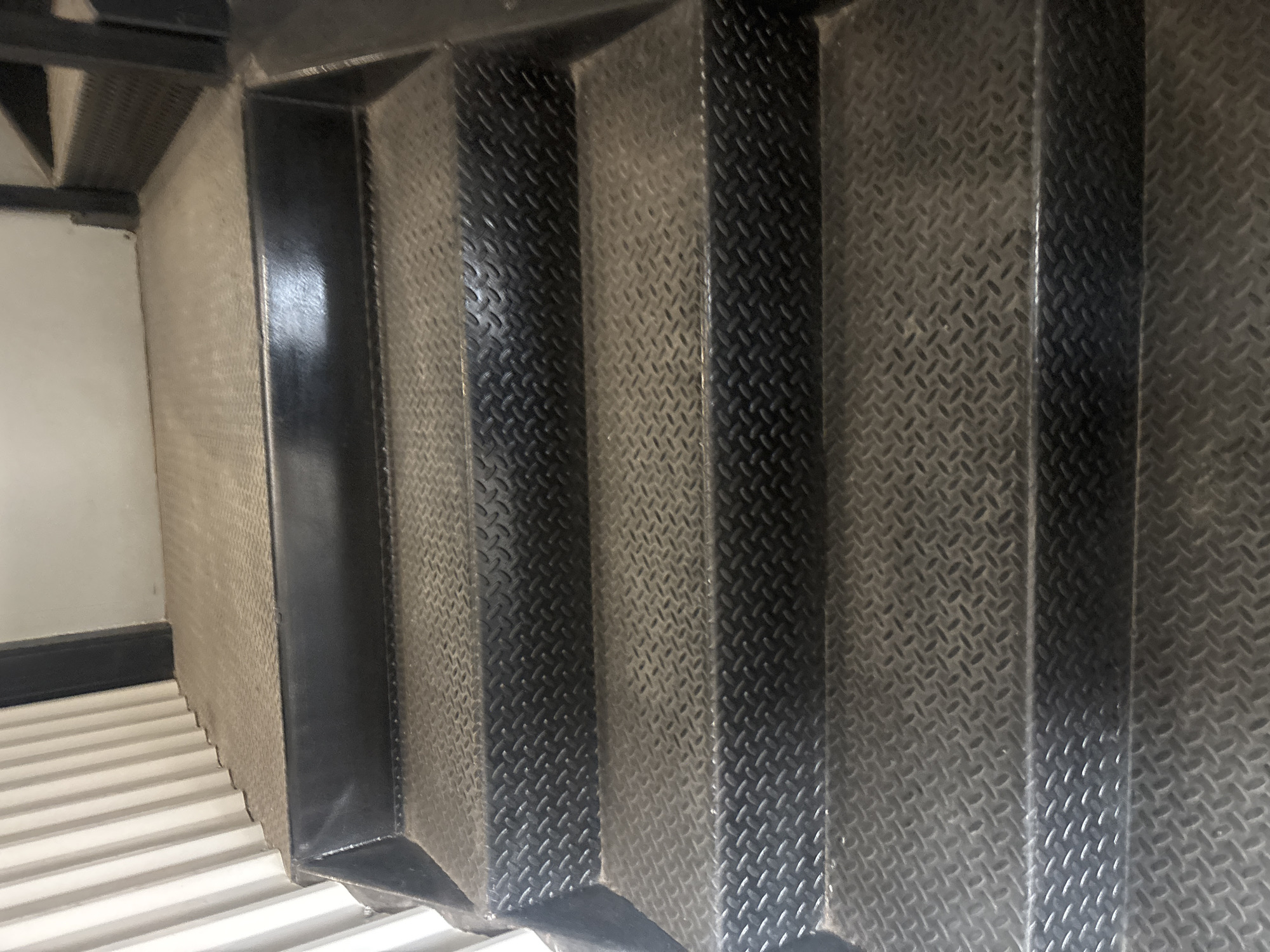 The image displays an upward-looking perspective of a set of industrial-style stairs. The steps are constructed from diamond plate metal, featuring a raised, repetitive pattern that provides an anti-slip surface. These step treads are primarily dark gray, with variations in shade due to lighting and wear. The risers and the structural supports at the edges of the steps are made of smooth, dark, possibly black, painted metal, reflecting light prominently.
The staircase is positioned centrally, ascending towards the top and right of the frame. On the left side of the stairs, there is a wall or supporting structure with a lighter, textured surface, possibly concrete or painted plaster, creating a contrast with the dark metal. Below the first visible step on the left, a section of a white, ribbed surface, potentially a floor or another architectural element, is visible.
No people are present, and no activity is occurring in the static scene. The lighting appears to be artificial, highlighting the metallic textures and creating subtle glints on the smooth surfaces. The overall aesthetic is functional and robust, suggesting a setting like a factory, warehouse, or a utility area within a larger building. There is no visible text or explicit location cues within the image itself.