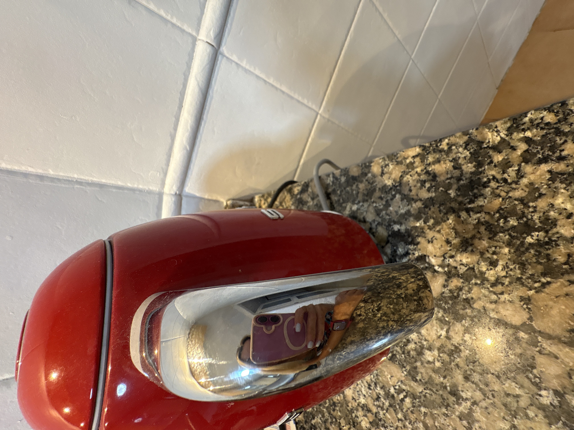 In València, Spain, a close-up shot shows a bright red kettle with a chrome handle. The kettle is positioned on a speckled granite countertop, with a white tiled wall in the background. A reflection in the chrome handle reveals a person holding a pink smartphone, taking a selfie. The phone case is pink and has a circular design with two cameras on the back. The person's hand, with white nail polish, is visible. A power cord for the kettle is seen plugged in, emerging from the base of the appliance. The lighting suggests an indoor setting, possibly a kitchen.