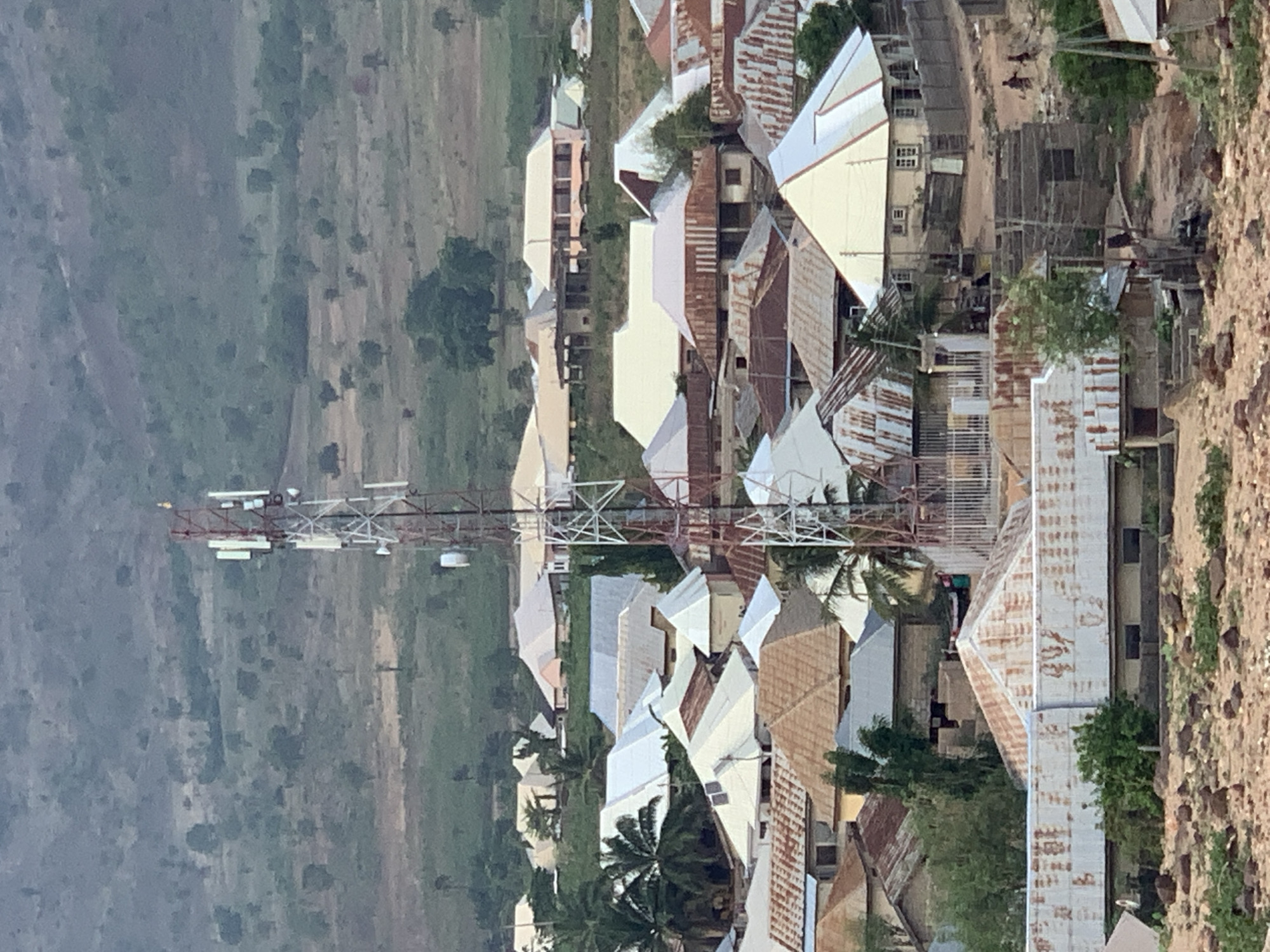 The image displays an elevated view of a settlement, likely a village or town, situated amidst a hilly and vegetated landscape. The scene is dominated by numerous buildings and a prominent telecommunications tower, under daylight conditions.
The most striking feature is a tall, red-and-white lattice telecommunications tower, equipped with multiple antennas and dishes. It stands centrally, towering over the surrounding structures. Clustered around the tower and extending across the midground are numerous buildings, predominantly with corrugated metal roofs. These roofs exhibit a range of appearances, from bright, reflective silver to heavily rusted reddish-brown, indicating varying ages and conditions. Some buildings are multi-story, while others appear to be single-level dwellings, with visible windows on some. Green trees and palm-like vegetation are interspersed among the buildings, adding natural elements to the built environment.
In the background, sweeping green hills or mountains are visible, covered in dense vegetation, suggesting a rural or mountainous setting. The ground closer to the buildings on the right side of the image appears to be rough, dry earth with scattered rocks. Two small, indistinct figures, possibly people, can be seen on a dirt path in the settlement, suggesting human activity, though no specific event is discernible.
The lighting indicates it is daytime, possibly late morning or afternoon, with no harsh shadows, suggesting diffused light. The overall scene provides a snapshot of a community in Lokoja, Nigeria, blending traditional housing with modern infrastructure within its natural environment. No visible text is present in the image.