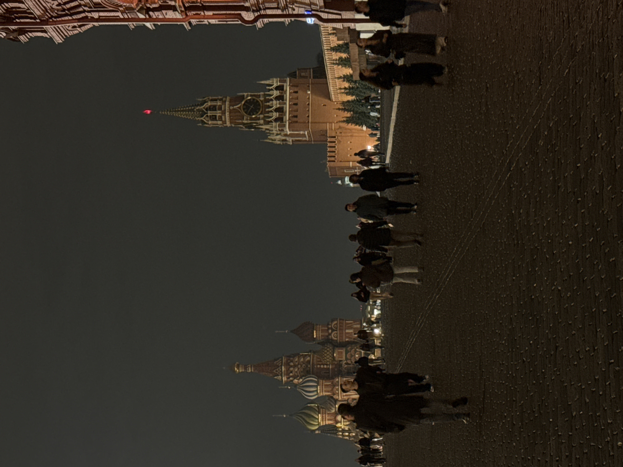 Captured at night, the image presents a view of Red Square in Moscow, Russia. Several individuals are present, some walking and others appearing to be standing or observing. The architectural landmarks of Saint Basil's Cathedral and the Spasskaya Tower, part of the Kremlin complex, are visible in the background, illuminated against the night sky. The ground, composed of cobblestone or paving, reflects some of the ambient light.