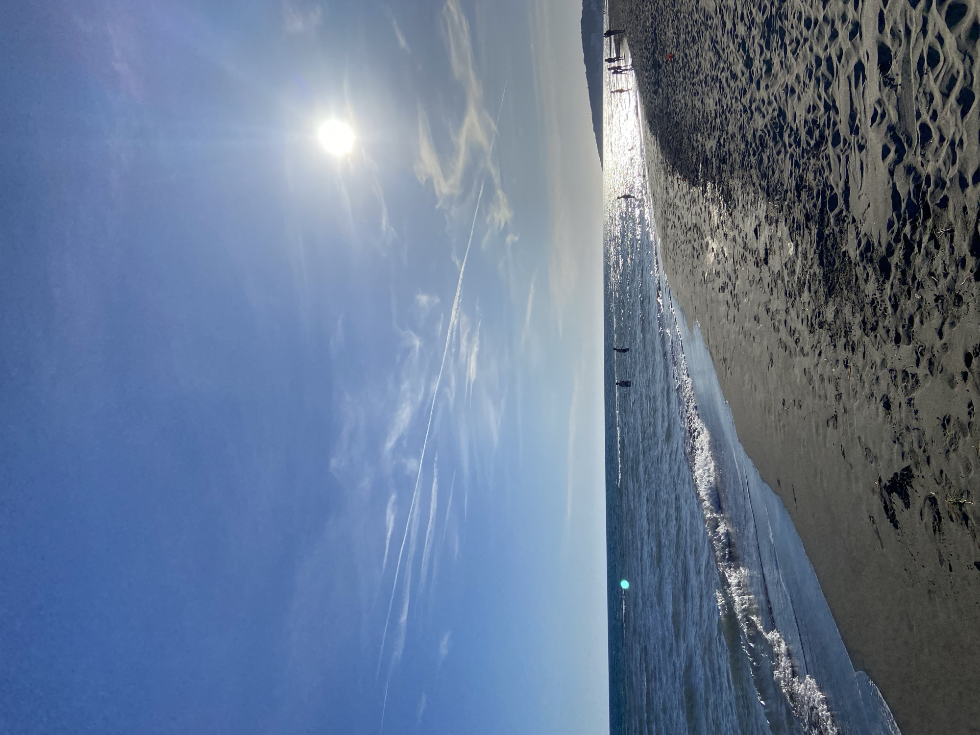 The image shows a beach scene. The sky is blue with some wispy clouds and contrails, and a bright sun. To the right of the sky is the ocean, with some visible waves. Along the edge of the ocean is a dark sand beach. There are a few distant figures on the beach and in the water. The city and specific location are unknown.