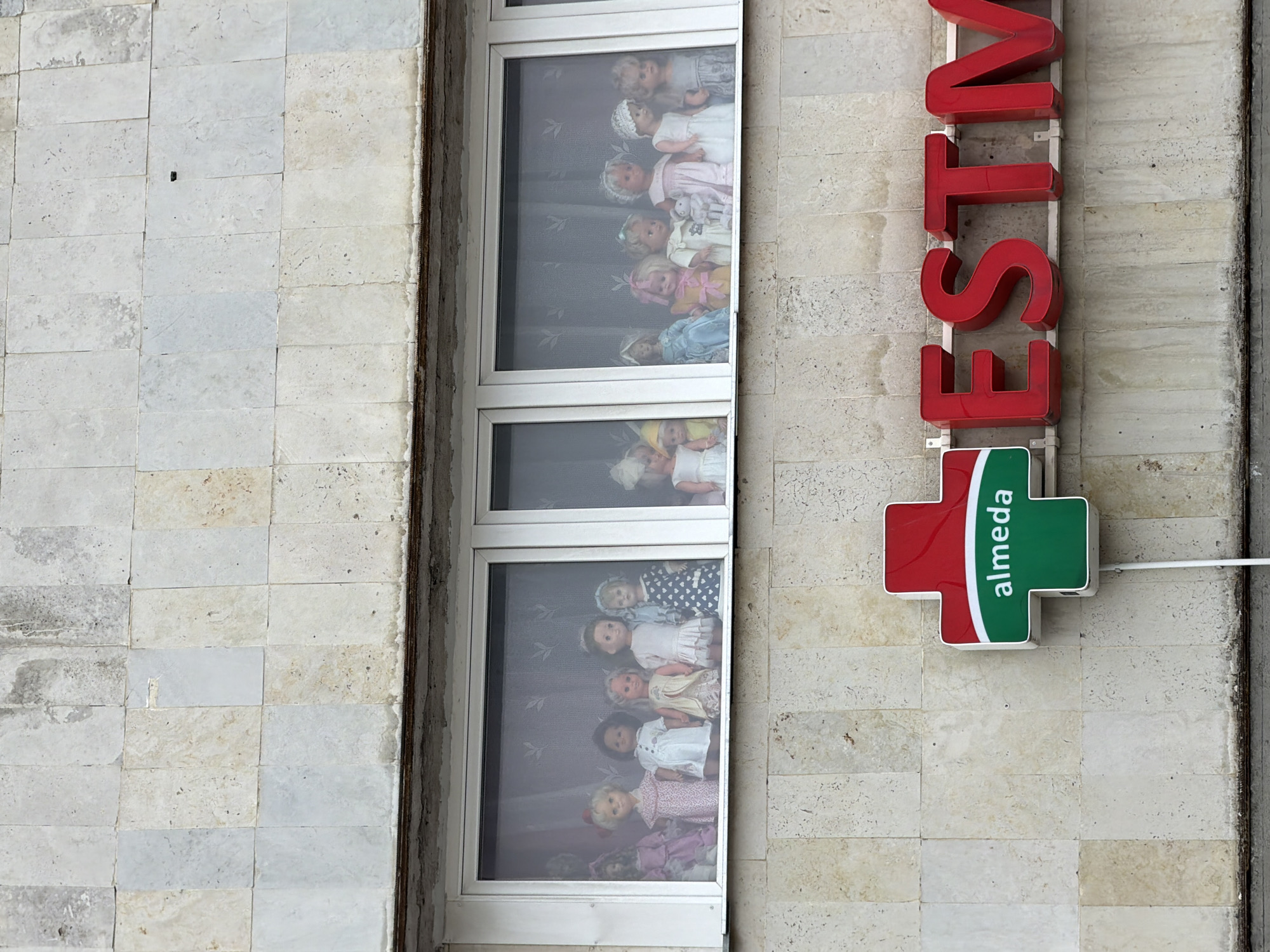 A vertical, medium shot captures the exterior of a building, likely a commercial establishment, in Tallinn, Estonia. The facade is constructed from rectangular, light-colored stone tiles. To the right of the image, a tall, red, three-dimensional sign spells out "ESTV" in block letters. Below the sign is a red and green cross emblem, a common symbol for pharmacies, with the word "almeda" written vertically in white on the green portion.
Adjacent to the sign are two windows. The upper window displays a collection of dolls arranged behind a sheer curtain. The dolls, appearing to be vintage, are dressed in various outfits and have light-colored hair. Some are wearing bonnets or caps. The lower window also shows a cluster of dolls, some with darker hair, also dressed in clothing and positioned behind a similar curtain. The dolls are arranged in a playful, almost life-like manner, as if sitting or standing.
The lighting suggests it is daytime, but the weather is not discernible. The scene is static, with no discernible activity. The overall impression is one of a quaint, perhaps older, storefront that has been decorated with a collection of dolls, possibly as a display or personal touch.