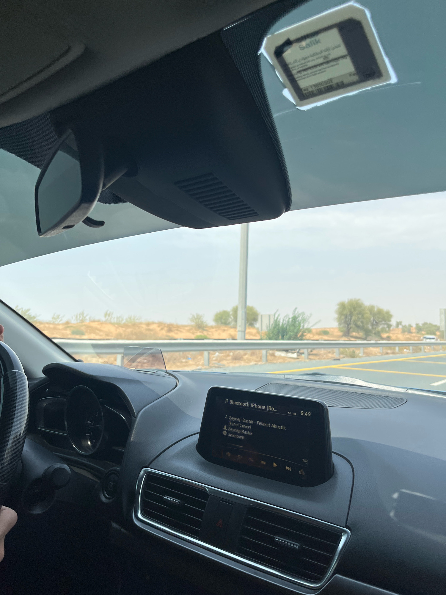 A driver's perspective shot from inside a car, looking forward. The dashboard is visible, with a screen displaying song information from a Bluetooth connection to an iPhone. The car is on a highway, with a landscape of desert vegetation and sandy hills visible through the windshield. A toll sticker is affixed to the windshield. The sky is overcast. The location is Mohadhub, United Arab Emirates.