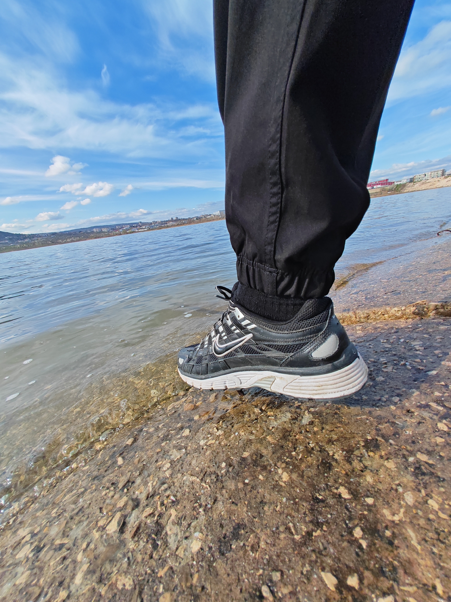 A close-up shot depicts a person standing on a stony shoreline next to a body of water. The person is wearing black pants gathered at the ankle and a black sneaker with white accents. The sneaker's sole is white and layered. The water is clear, with small waves lapping against the shore. The shoreline consists of small stones and pebbles. In the background, across the water, a cityscape is visible under a blue sky with scattered clouds. The city in the background is Shumen, Bulgaria.