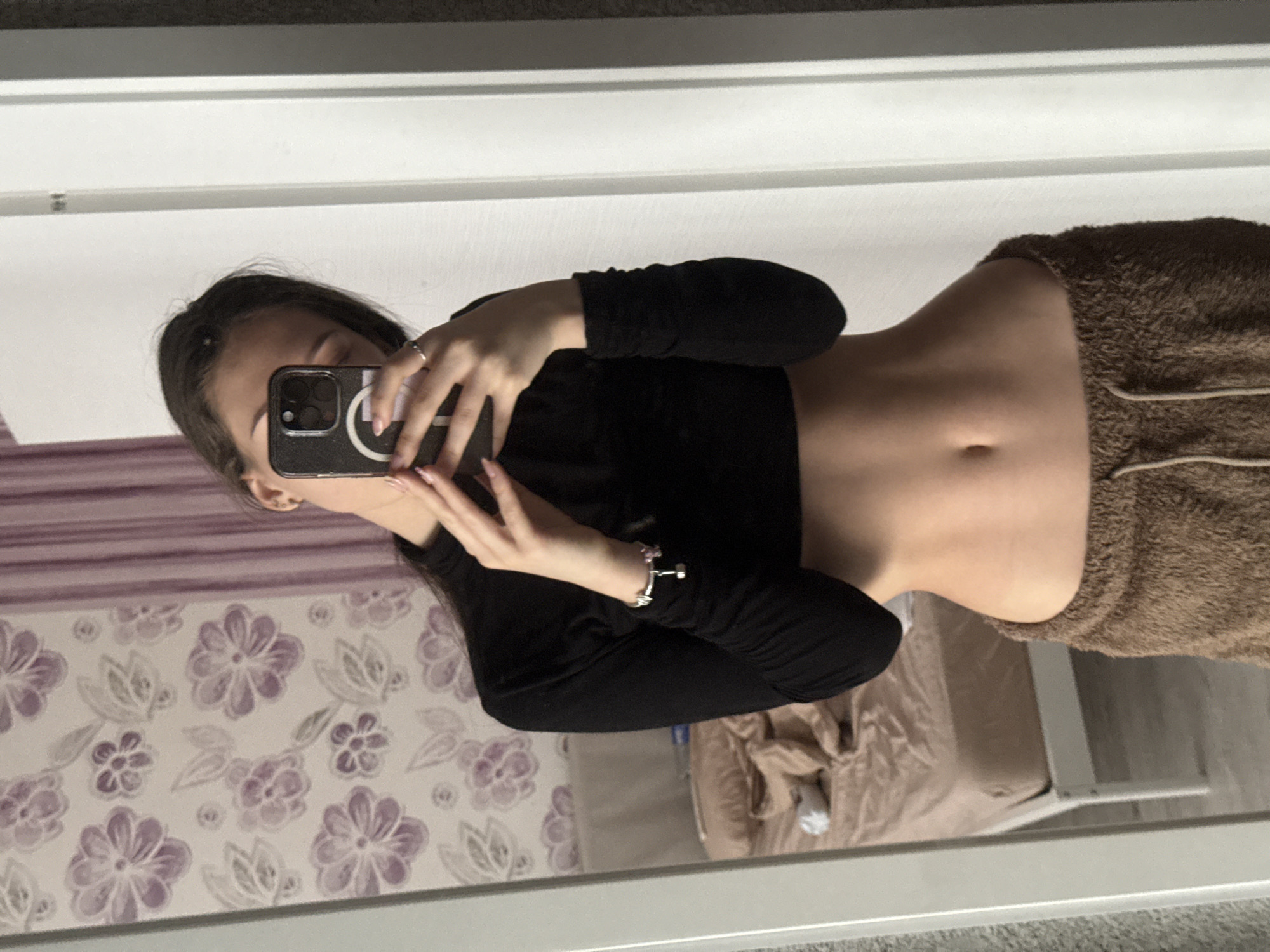 A mirror selfie shows a young woman with long, dark hair, wearing a black long-sleeved cropped shirt and brown shorts. She is holding her phone to take the picture, partially obscuring her face. Her stomach is visible. The background is blurred and shows a bedroom with floral wallpaper, a bed with tan bedding, and a white frame suggesting a mirror or a window. There is no indication of the location being Sofia, Bulgaria.
