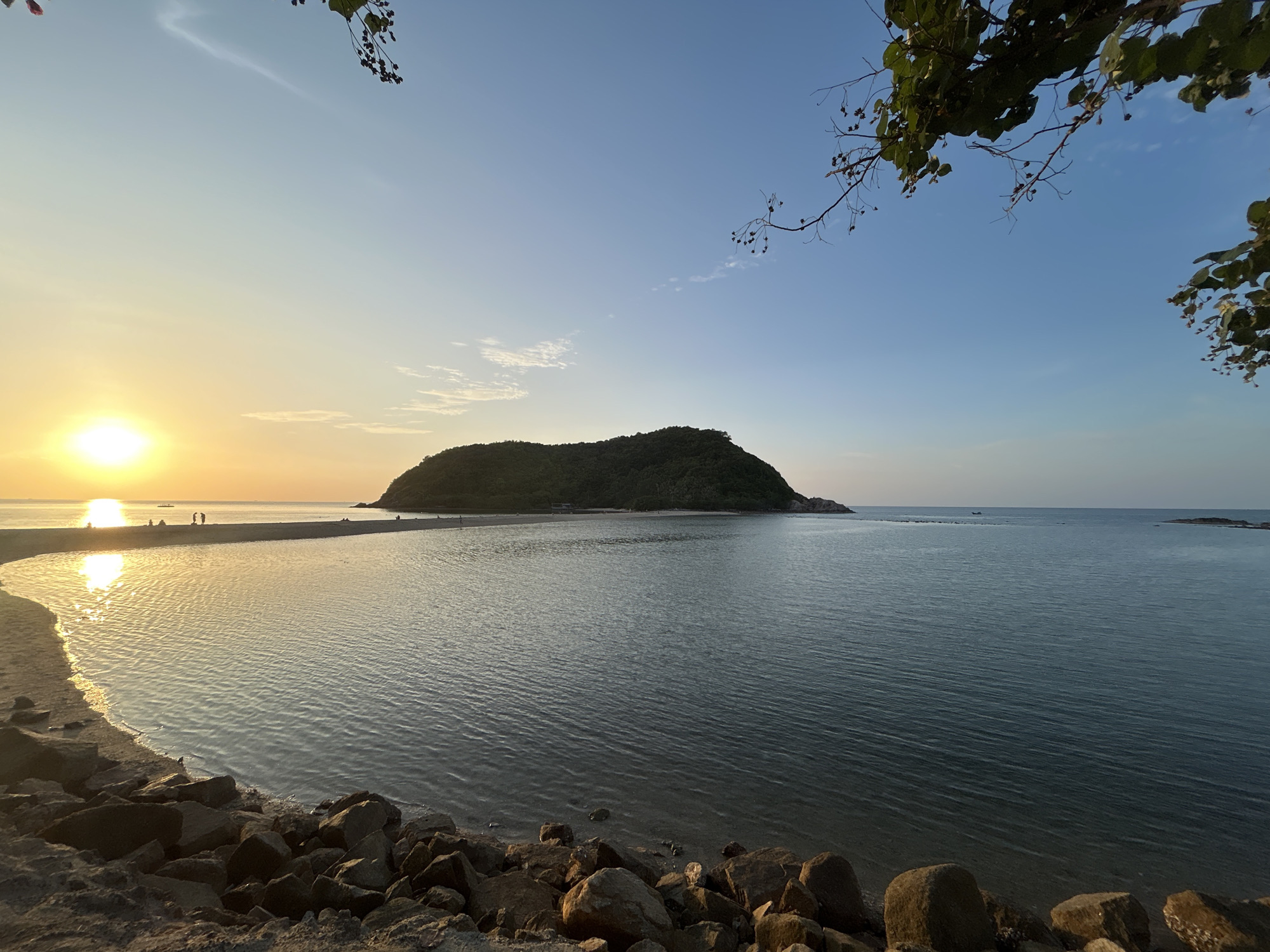 A scenic view of a tranquil beach on Ko Pha-ngan District, Thailand, captures the serene beauty of a sunset over the ocean. The sun, a bright orb of light, casts a warm glow across the sky, reflecting on the calm water. A narrow strip of sand connects the beach to a lush, green island in the distance. A few people are visible on the sandbar, silhouetted against the radiant sun. The foreground is adorned with a scattering of rocks, adding a touch of natural texture to the composition. The sky is a vast expanse of blue, with wispy clouds adding depth and character. The overall ambiance is one of peacefulness and natural splendor, inviting viewers to immerse themselves in the beauty of this tropical paradise.