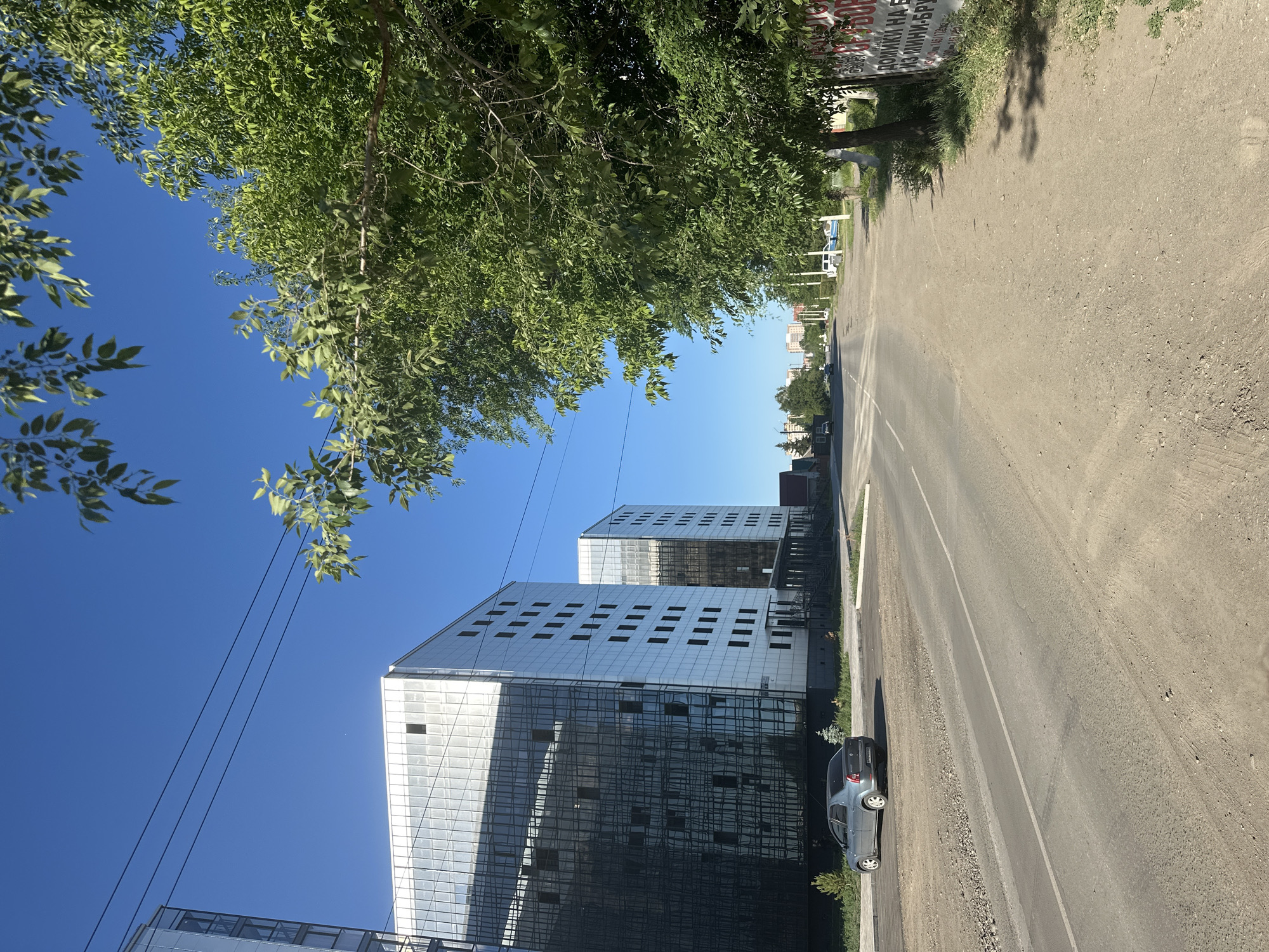 The image depicts a street scene in Kalinin, Russia, under a clear, bright blue sky, suggesting a sunny day. Lush green trees with dense foliage frame the top left of the image, casting dappled shadows. To the right, a paved road runs diagonally, with a gravel shoulder. Along the left side of the road, modern buildings with reflective glass facades dominate the scene. These multi-story structures have a contemporary architectural style, with numerous rectangular windows. A silver car is parked on the gravel shoulder near one of the buildings. Further down the road, more buildings are visible, some appearing residential. A large sign with red and white lettering is affixed to a tree on the right side of the road, though the text is partially obscured and difficult to read clearly. The overall impression is one of an urban or semi-urban environment on a pleasant day, with a contrast between the natural elements of the trees and the modern architecture.