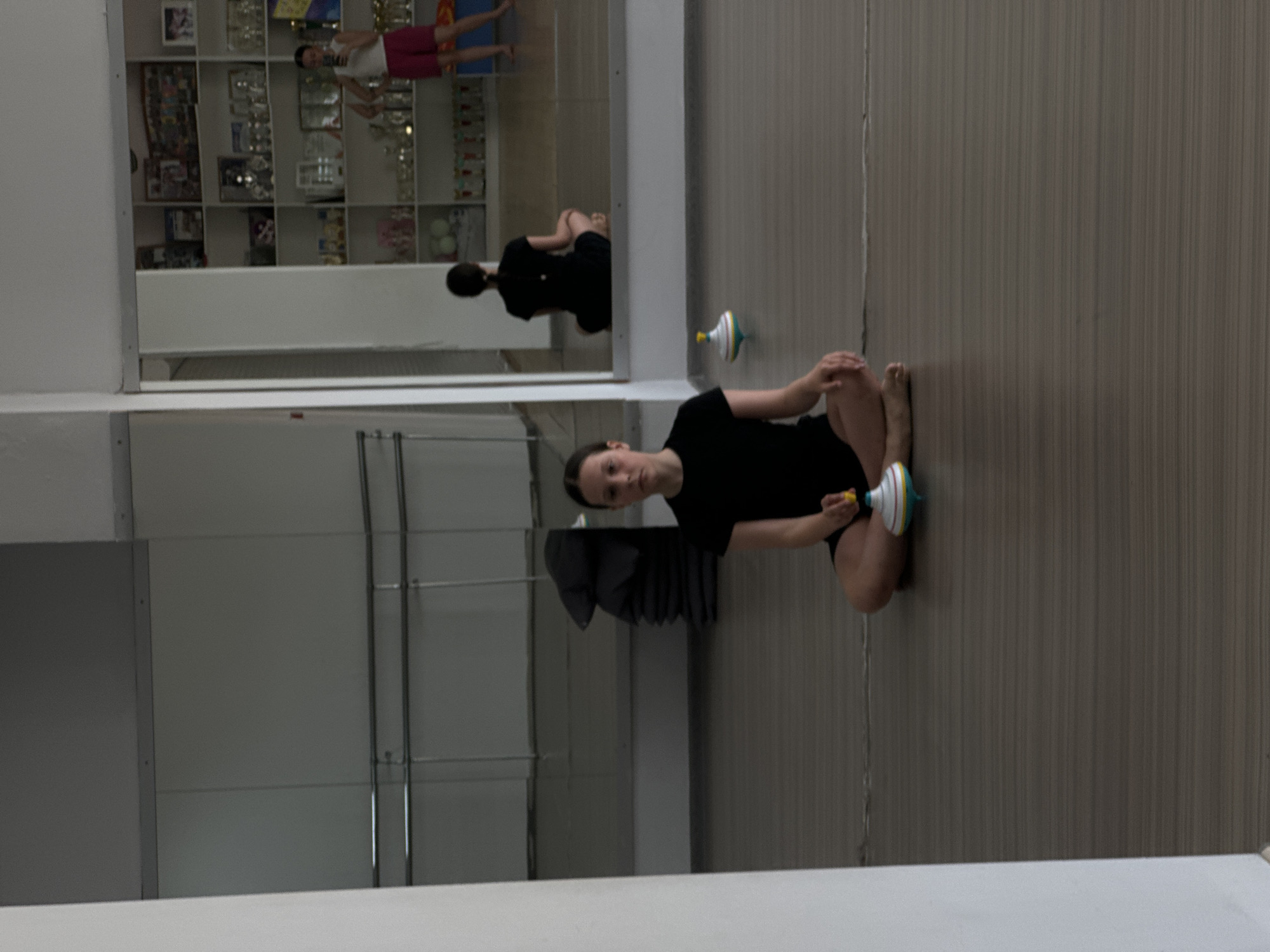 The scene is a dance studio in Votkinsk, Russia. A young woman wearing a black t-shirt and shorts is sitting cross-legged on the floor, holding a colorful spinning top. She is positioned in the center of the frame, facing the viewer. Behind her, there is a wall with horizontal panels and vertical support beams. To the left of the woman, there is a large mirror reflecting the studio. The reflection shows another view of the woman, as well as a shelf filled with trophies and other items in the background. The overall lighting is bright and diffused, suggesting it is daytime. The floor appears to be a light gray or beige color with vertical stripes.