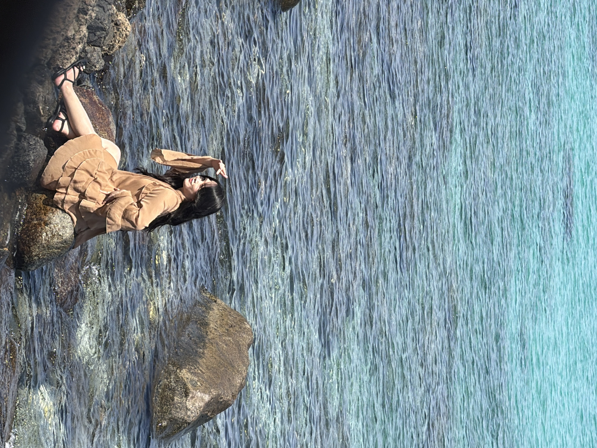A woman is lying on rocks by the water's edge in Nha Trang Ward, Vietnam. She is wearing a tan dress and black sandals. Her black hair flows down her back. The water is a mix of light blue and grey. The rocks are dark and smooth.