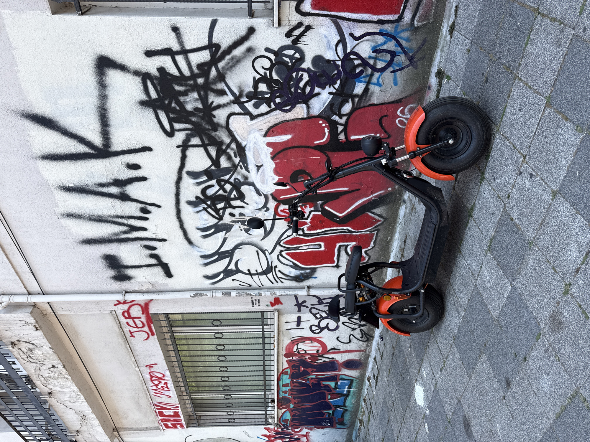 An orange and black electric scooter is parked on a gray tiled sidewalk in front of a white wall covered in graffiti. The wall features various tags and graffiti in black, red, and blue spray paint, including large block letters spelling "IMAK" and other illegible markings. A barred window is visible on the left side of the wall, with more graffiti beneath it. The scooter is positioned facing right, with its front wheel and fender prominently displayed. The environment appears to be an urban alleyway or street with a slightly worn appearance. The lighting suggests it is daytime.