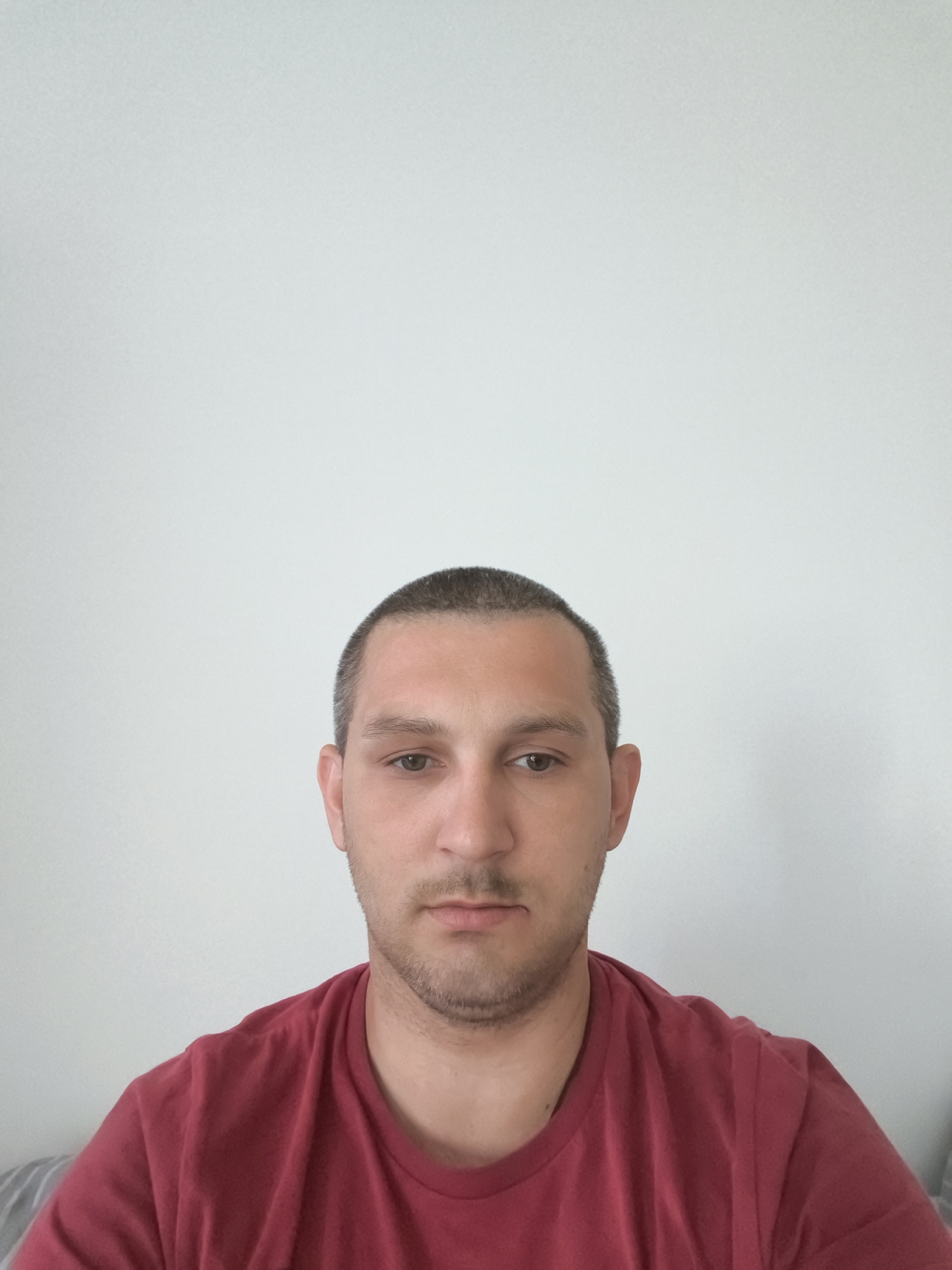 A young adult male with short brown hair and a stubbled beard looks directly at the camera. He is wearing a plain red t-shirt. The background is a plain, light-colored wall. The image is a head-and-shoulders shot, likely taken indoors in a residential setting. There is no information in the image to suggest the location is in Wolgast, Germany.