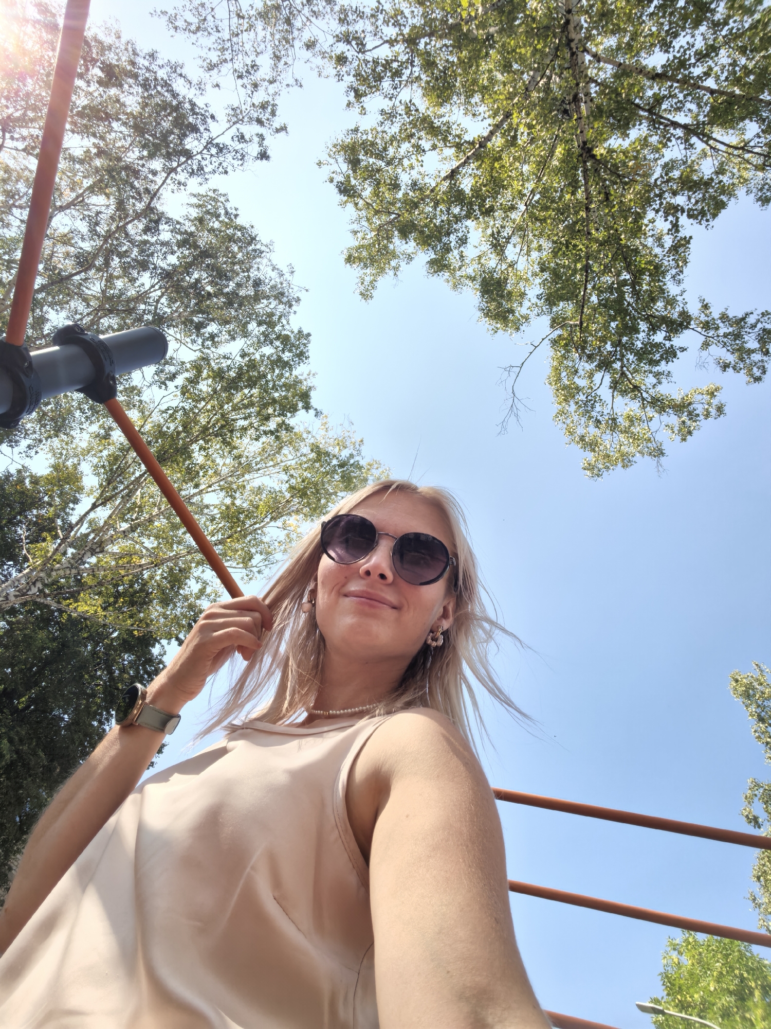 A young woman with blonde hair, wearing sunglasses, a light-colored top, and a pearl necklace, is captured in a selfie outdoors. She is holding her hair and looking towards the camera with a slight smile. The background consists of a clear blue sky and the leafy branches of trees, suggesting a sunny day. A dark-colored object, possibly part of a scooter or bicycle, is visible on the left side of the frame, along with orange bars that appear to be part of a railing or structure. The scene is set in the city of Sterlitamak, Russia, though no specific landmarks identifying the location are visible in the image.